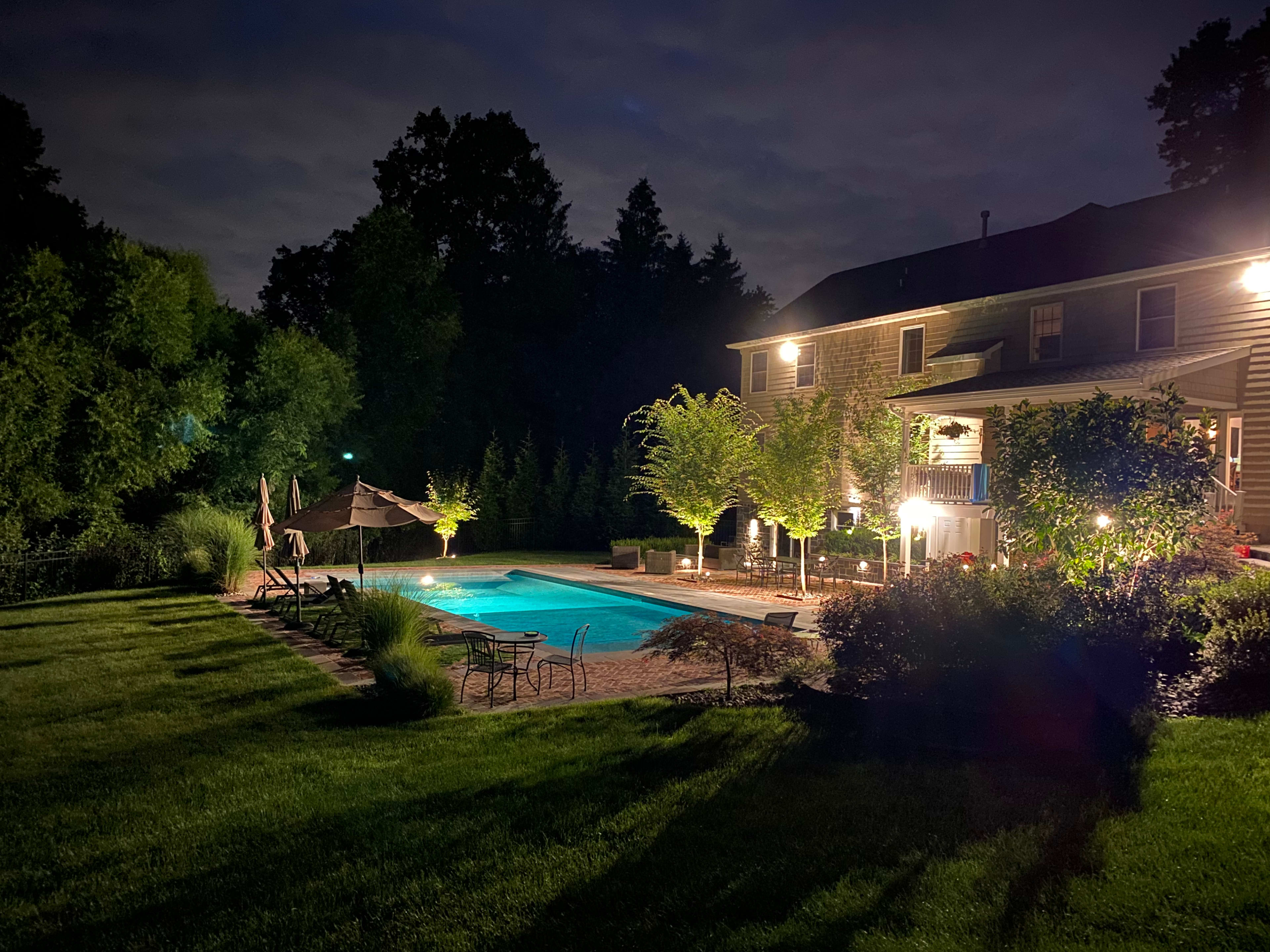 A well-lit backyard features a swimming pool surrounded by lounge chairs and landscaping, with a house visible in the background under a night sky.