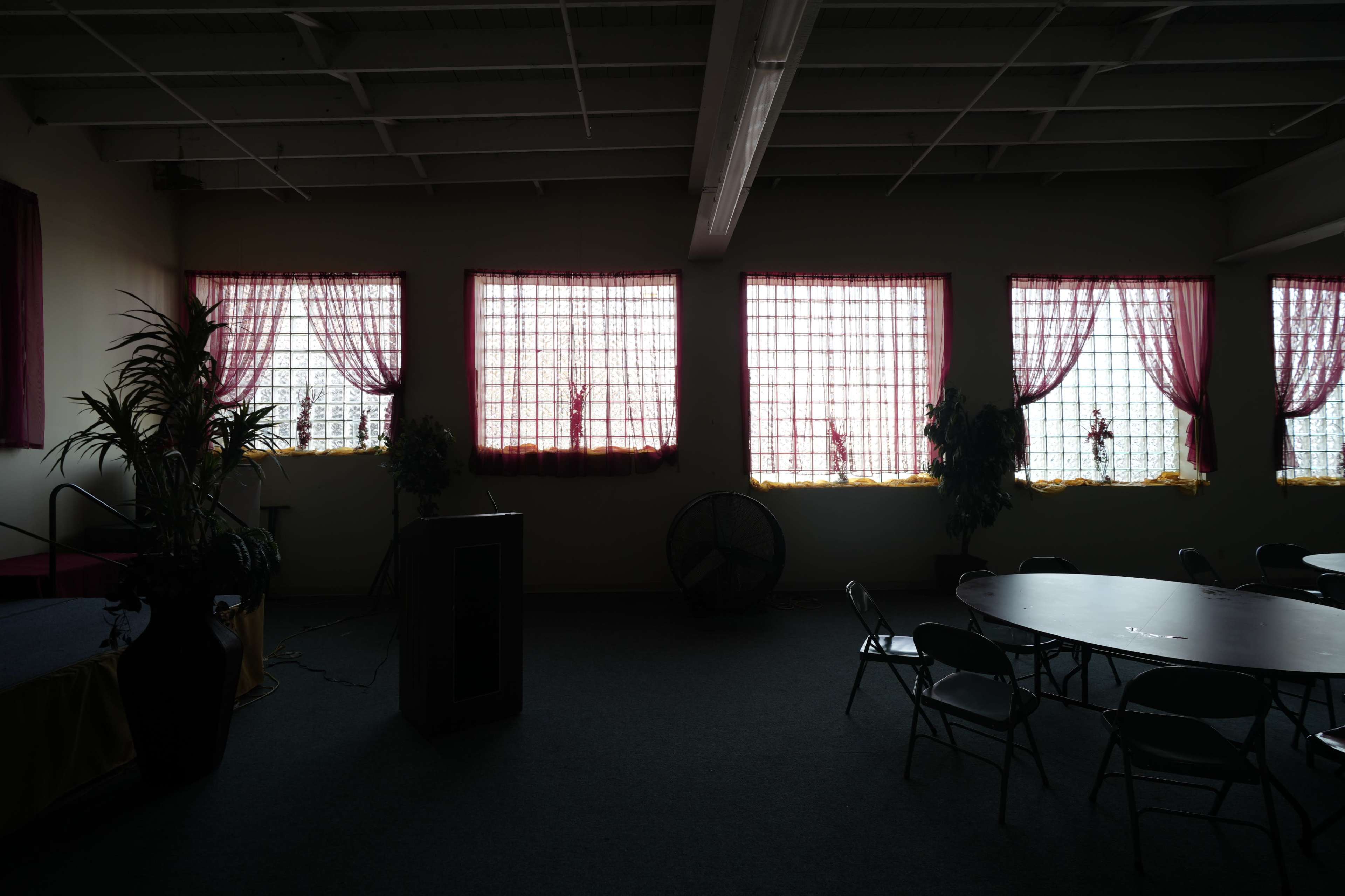 The image shows a dimly lit room with several windows covered by sheer curtains and a round table surrounded by chairs.