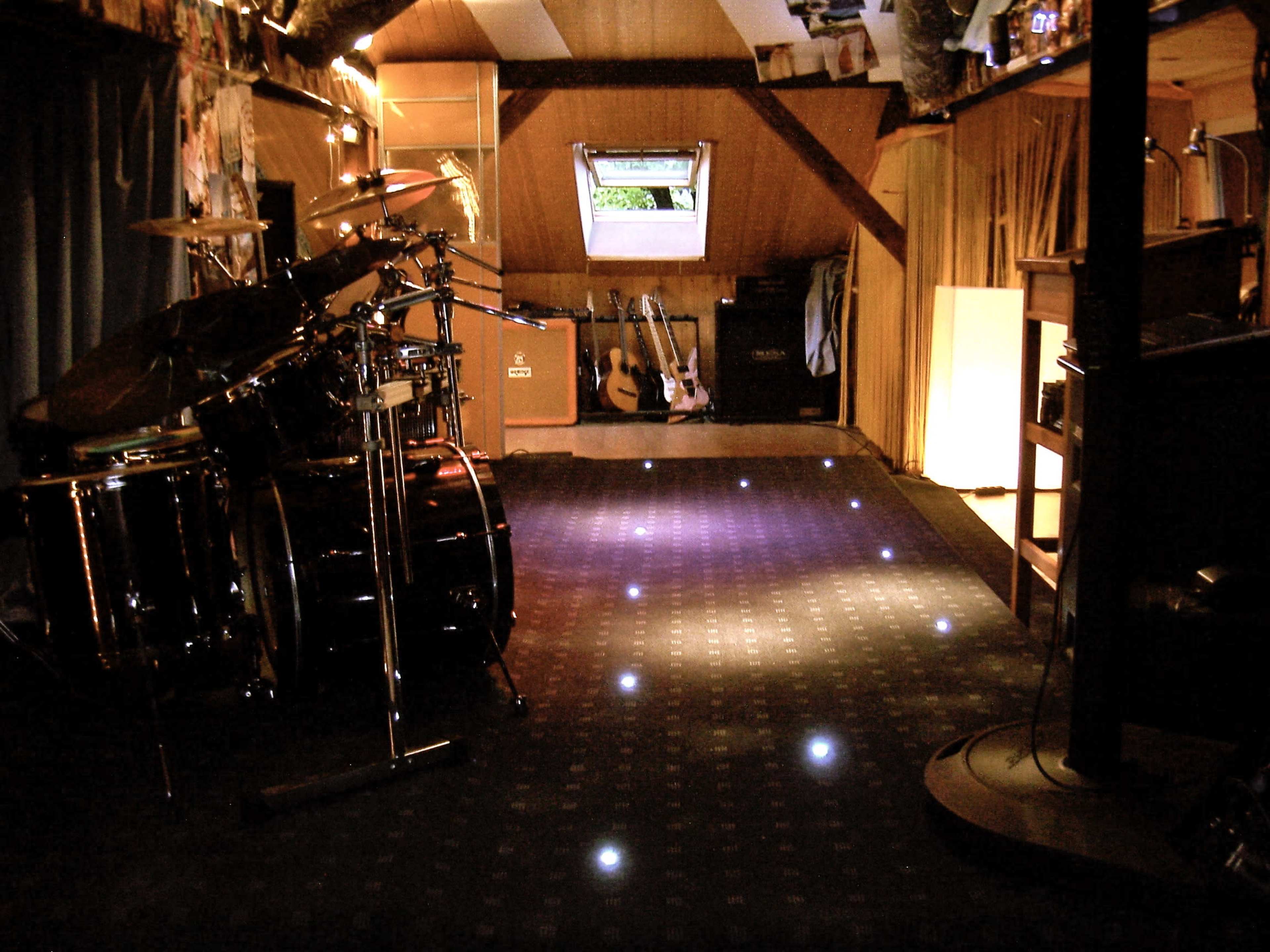 A dimly lit attic space set up as a music rehearsal room, featuring a drum kit, several guitars on stands, and a carpeted floor with small lights.