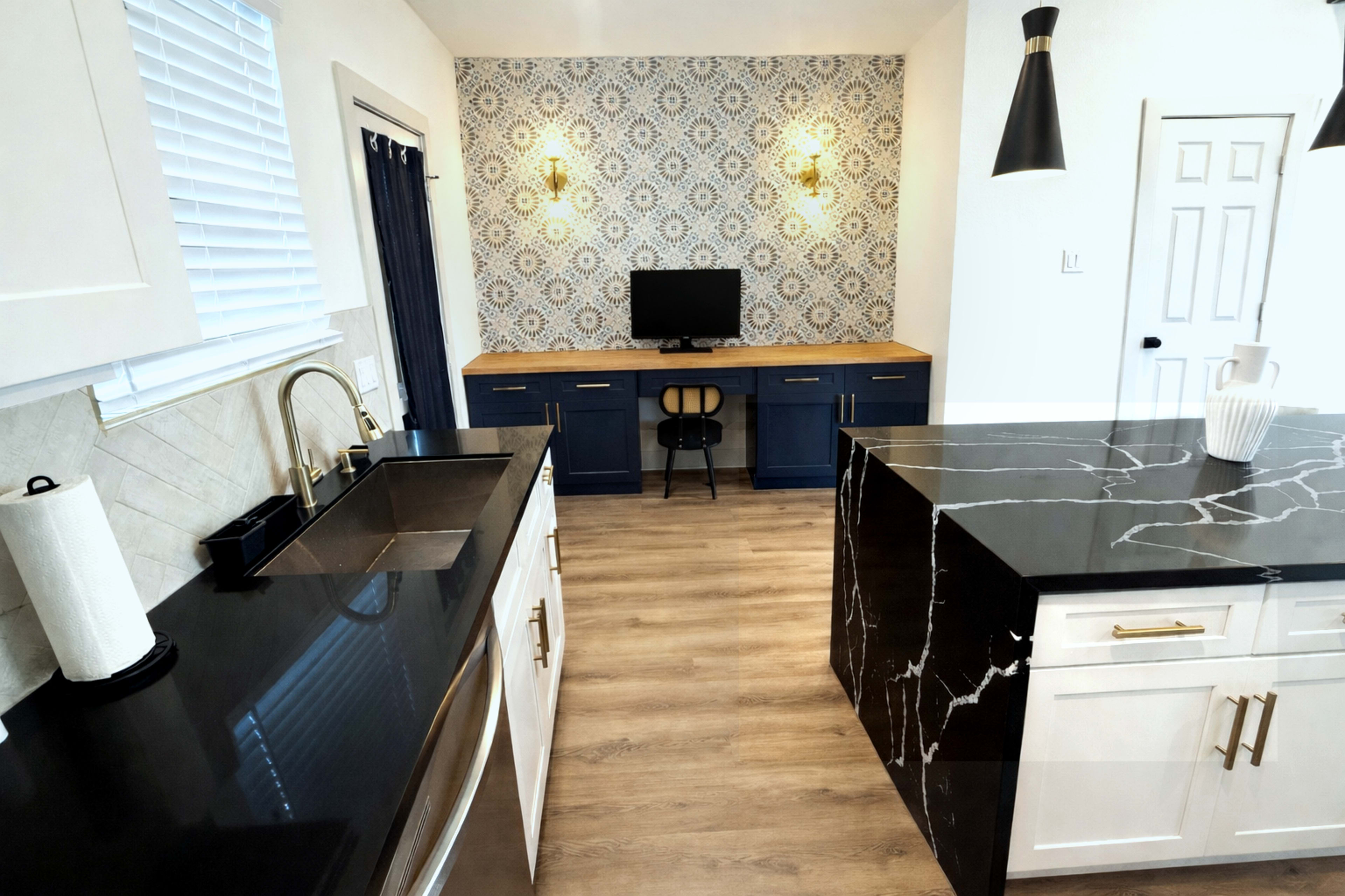 The image shows a modern kitchen with white cabinetry and black countertops, complemented by a patterned wall and a small desk area with a monitor.