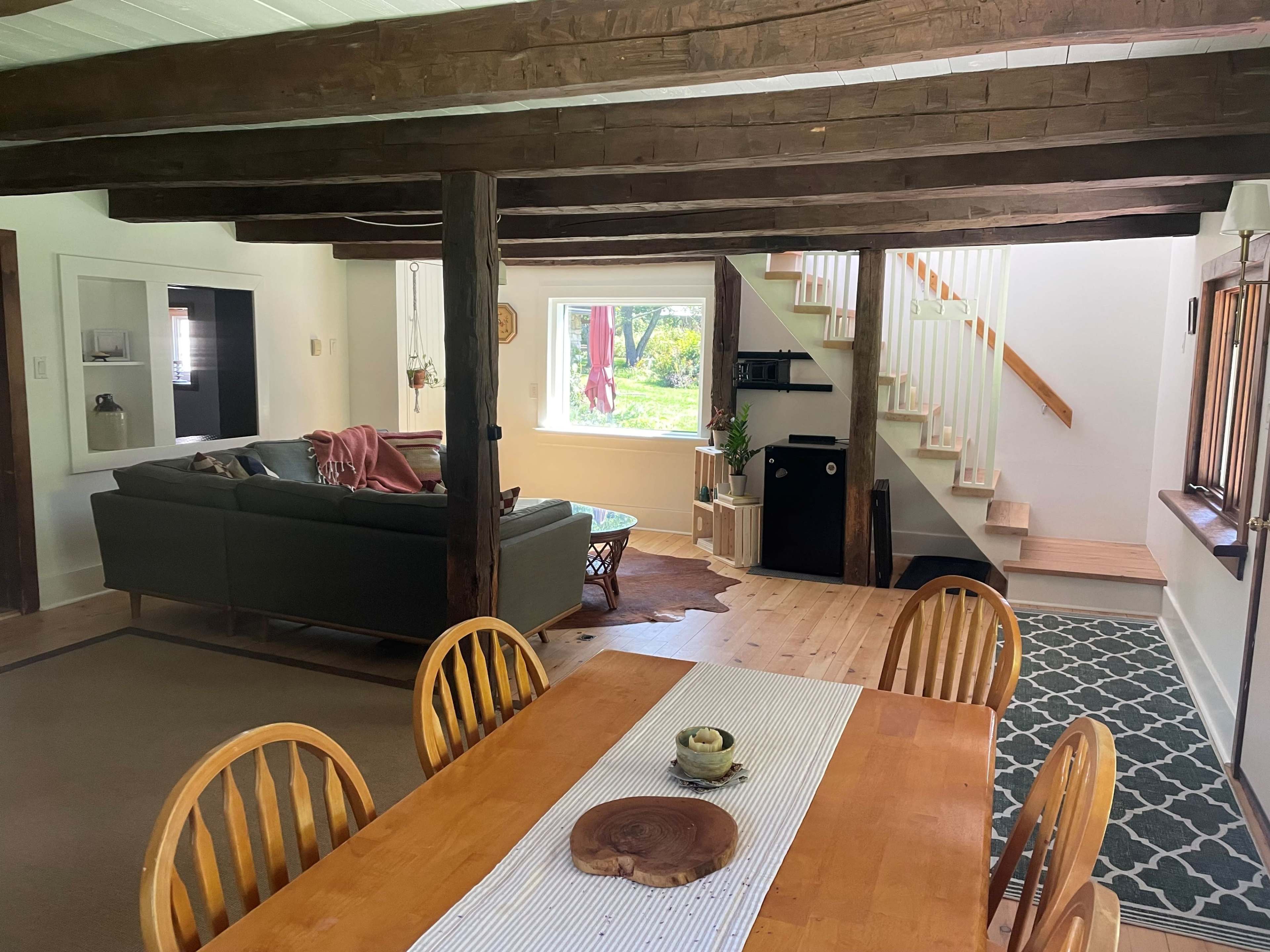 A living room and dining area feature wooden beams, a staircase, and large windows overlooking a green view.
