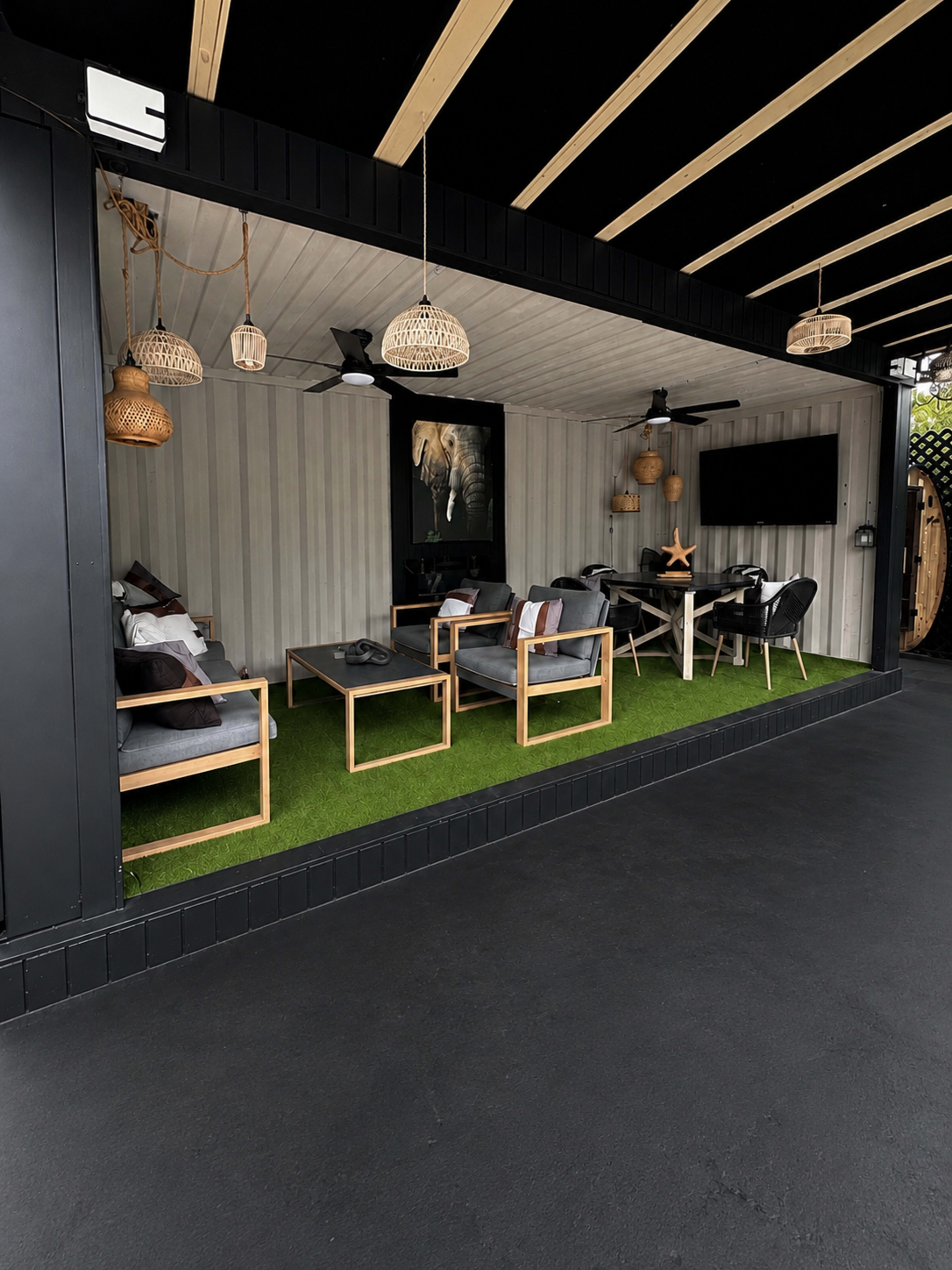 The image shows a covered outdoor seating area with modern furniture, a wall-mounted television, and decorative elements, all set against a backdrop of artificial grass.