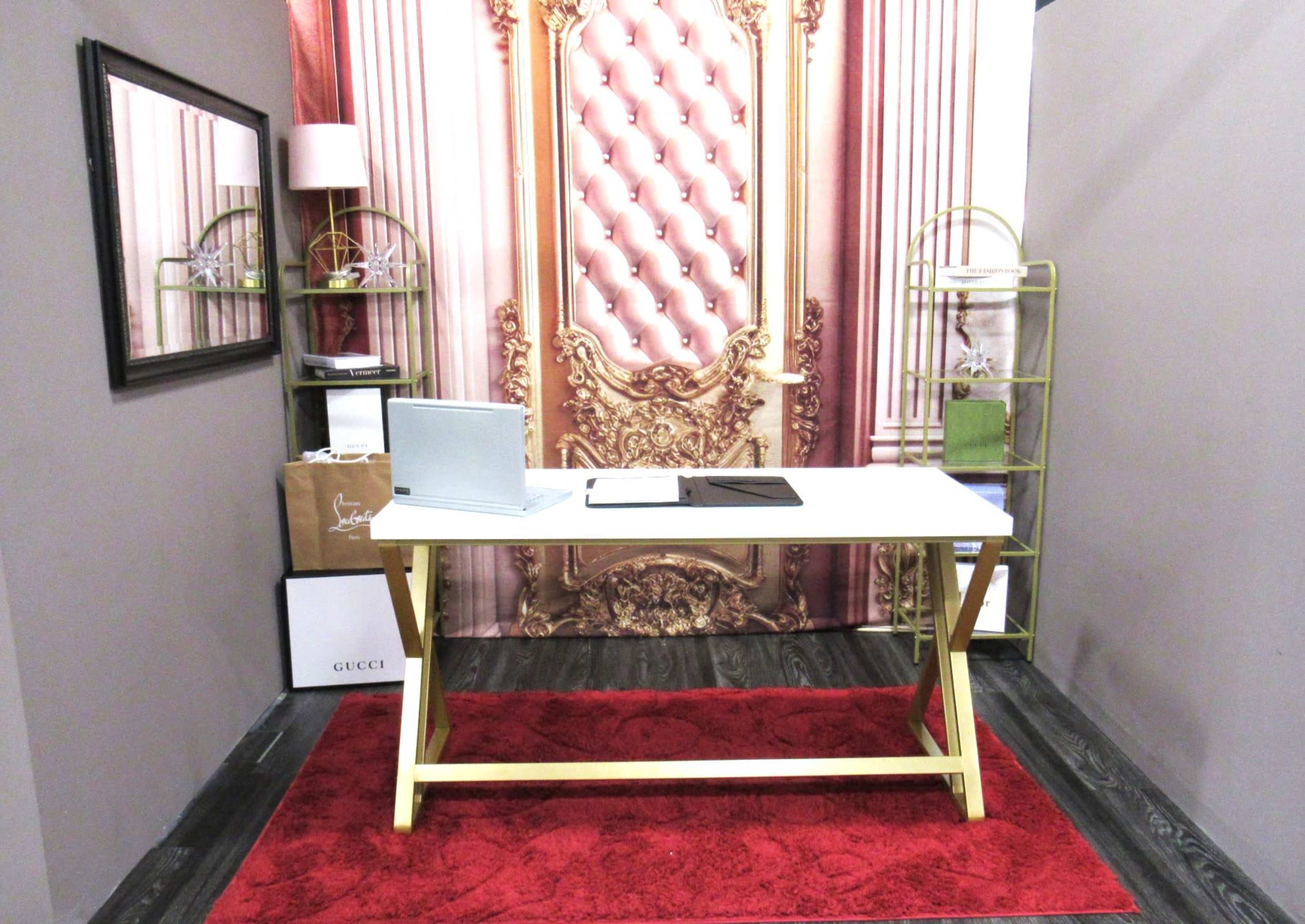 A stylish office space featuring a white desk with a laptop and a tablet, set against a backdrop of an ornate pink wallpaper and accented by golden shelves and a red rug.