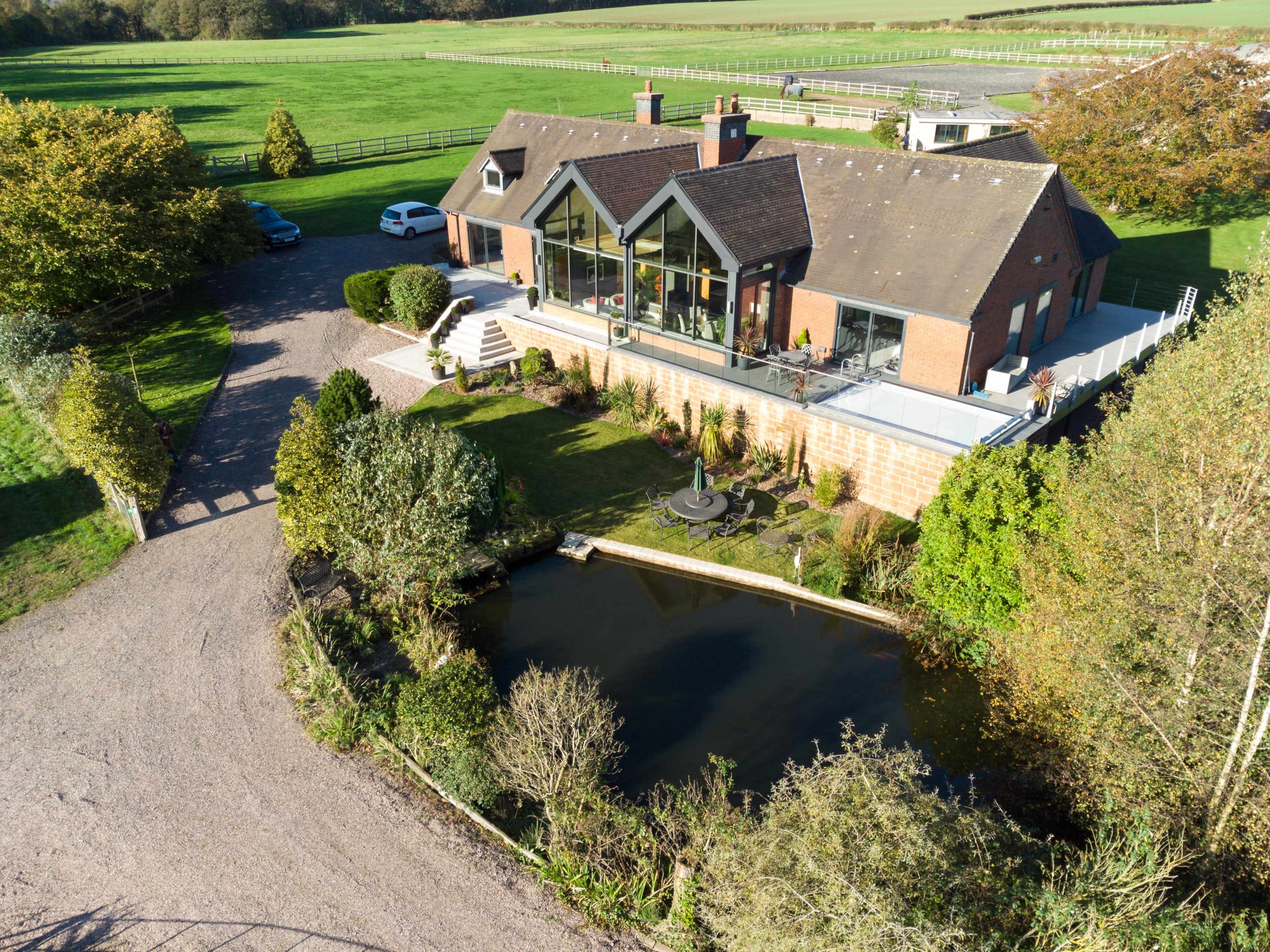 The image shows a large brick house with a modern glass extension, surrounded by greenery and a pond, situated on a landscaped property.