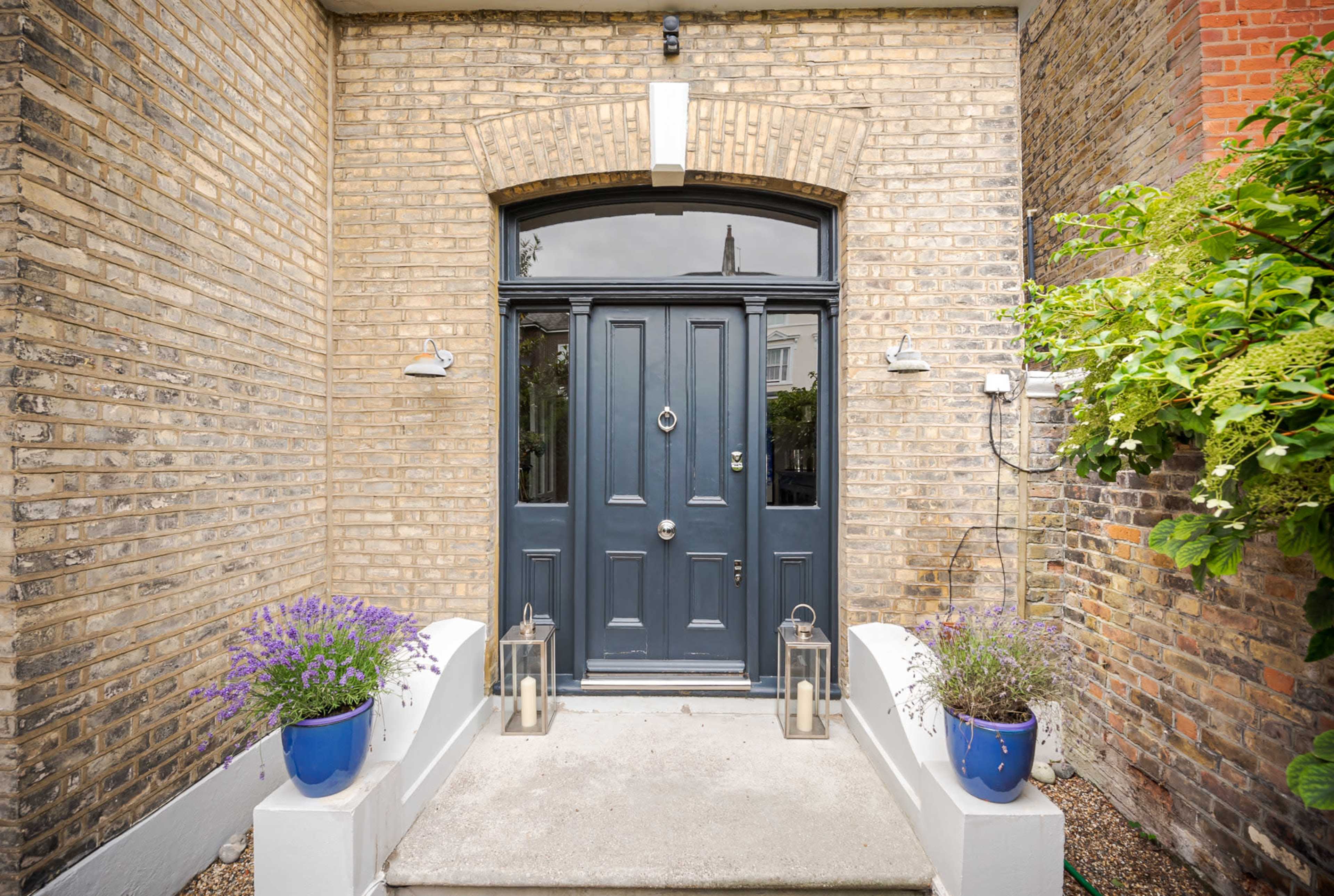 The image shows a dark blue front door set within a brick wall, flanked by purple flowers in pots and lanterns on either side.