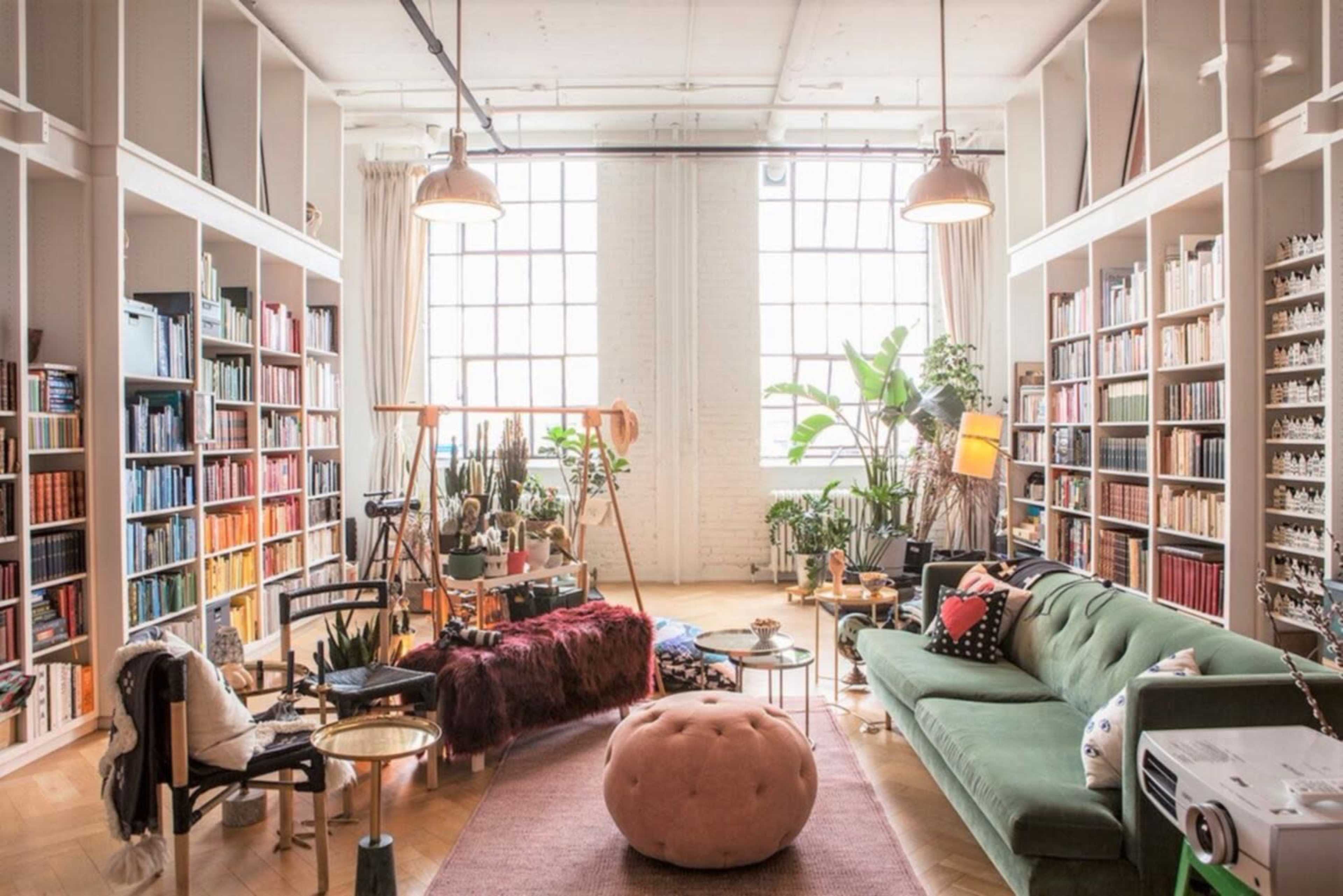 A spacious living room features large windows, bookshelves filled with colorful books, a green sofa, and various plants.