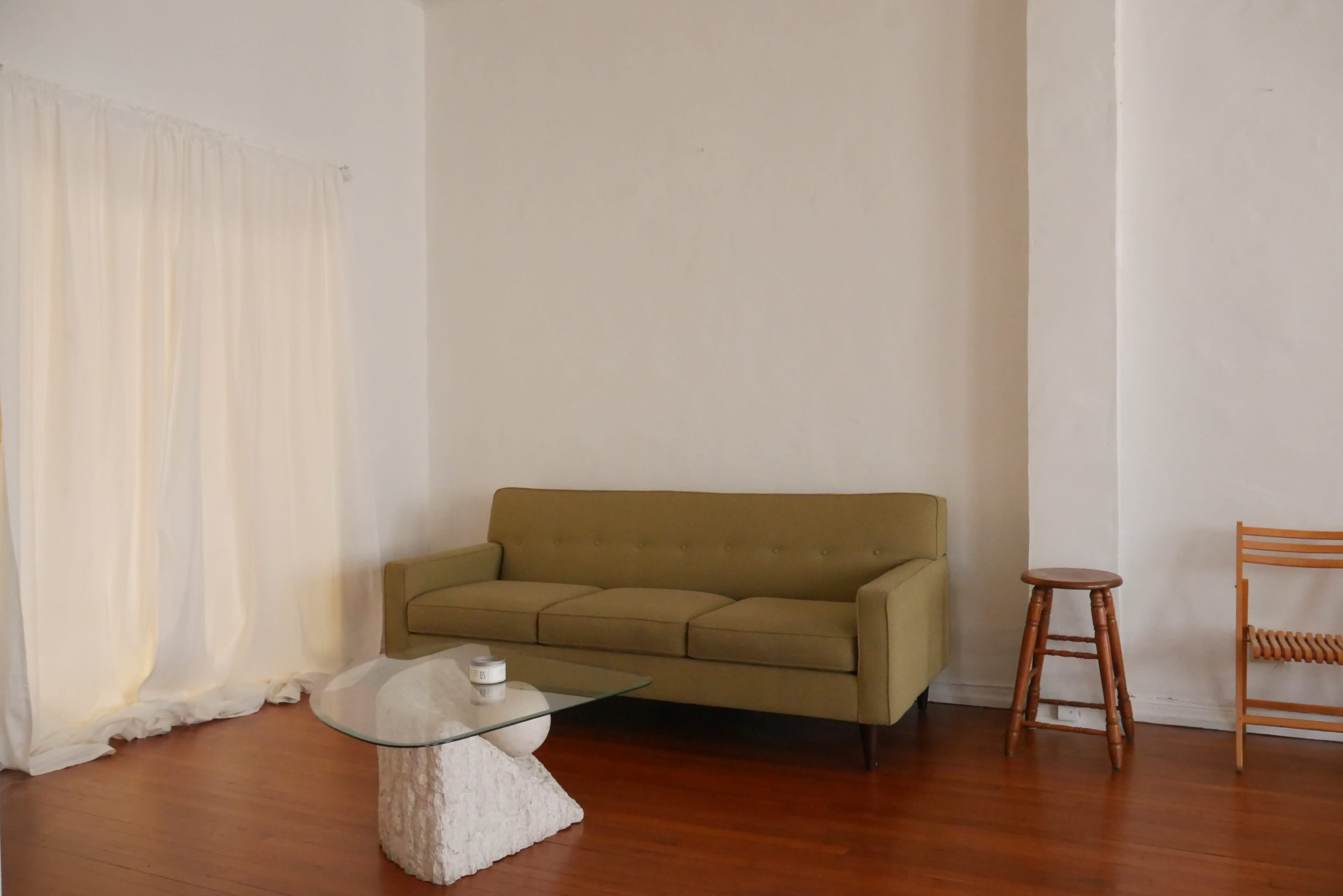 The image shows a minimalistic living room featuring a green sofa, a glass coffee table, a wooden stool, and sheer curtains by a window.
