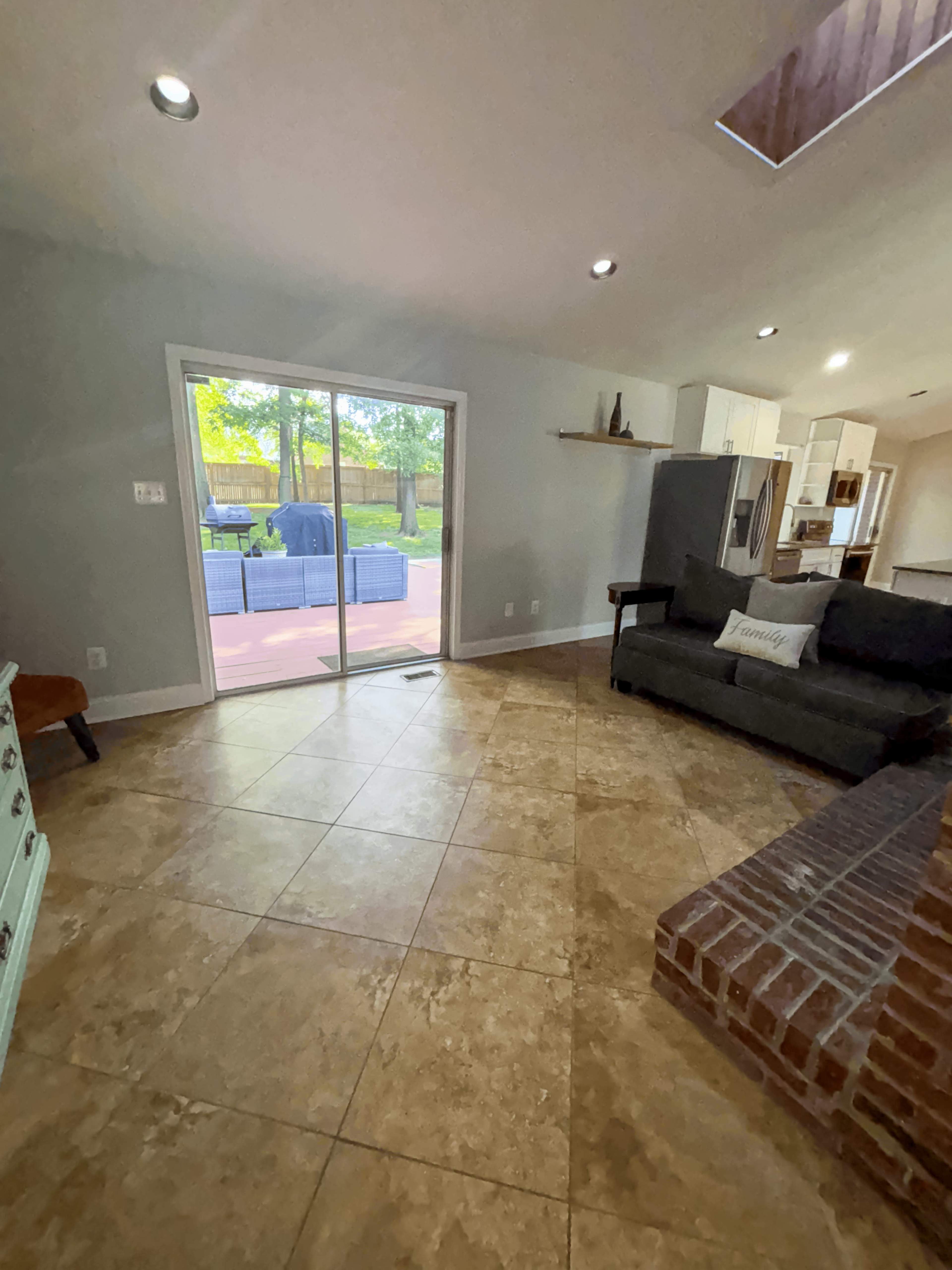 The image shows a living area with tile flooring, a dark sofa, and a sliding glass door leading to an outdoor patio.