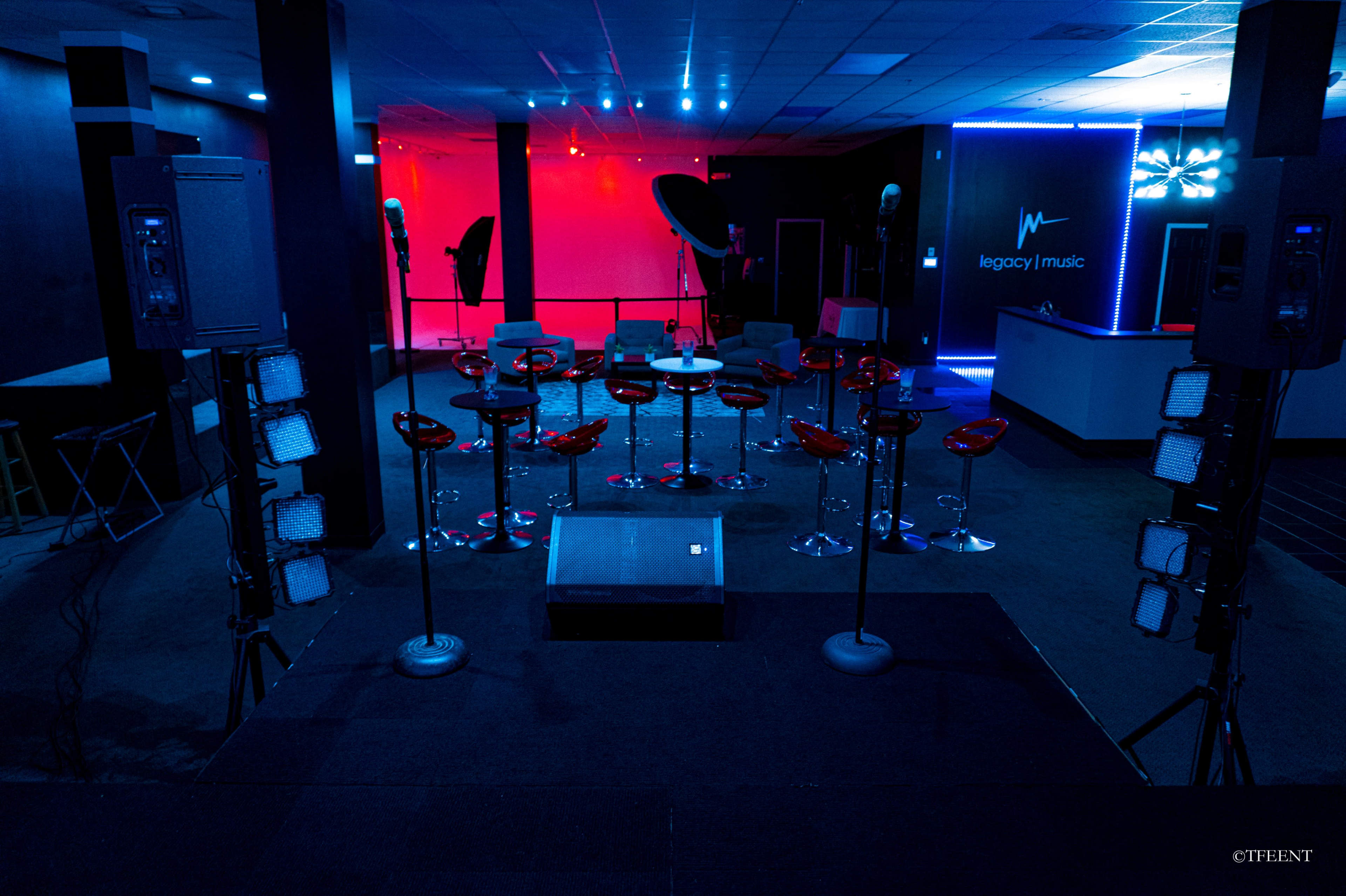 A dimly lit music venue featuring a stage with a sound system, illuminated red and blue lights, and a row of red stools facing the stage.