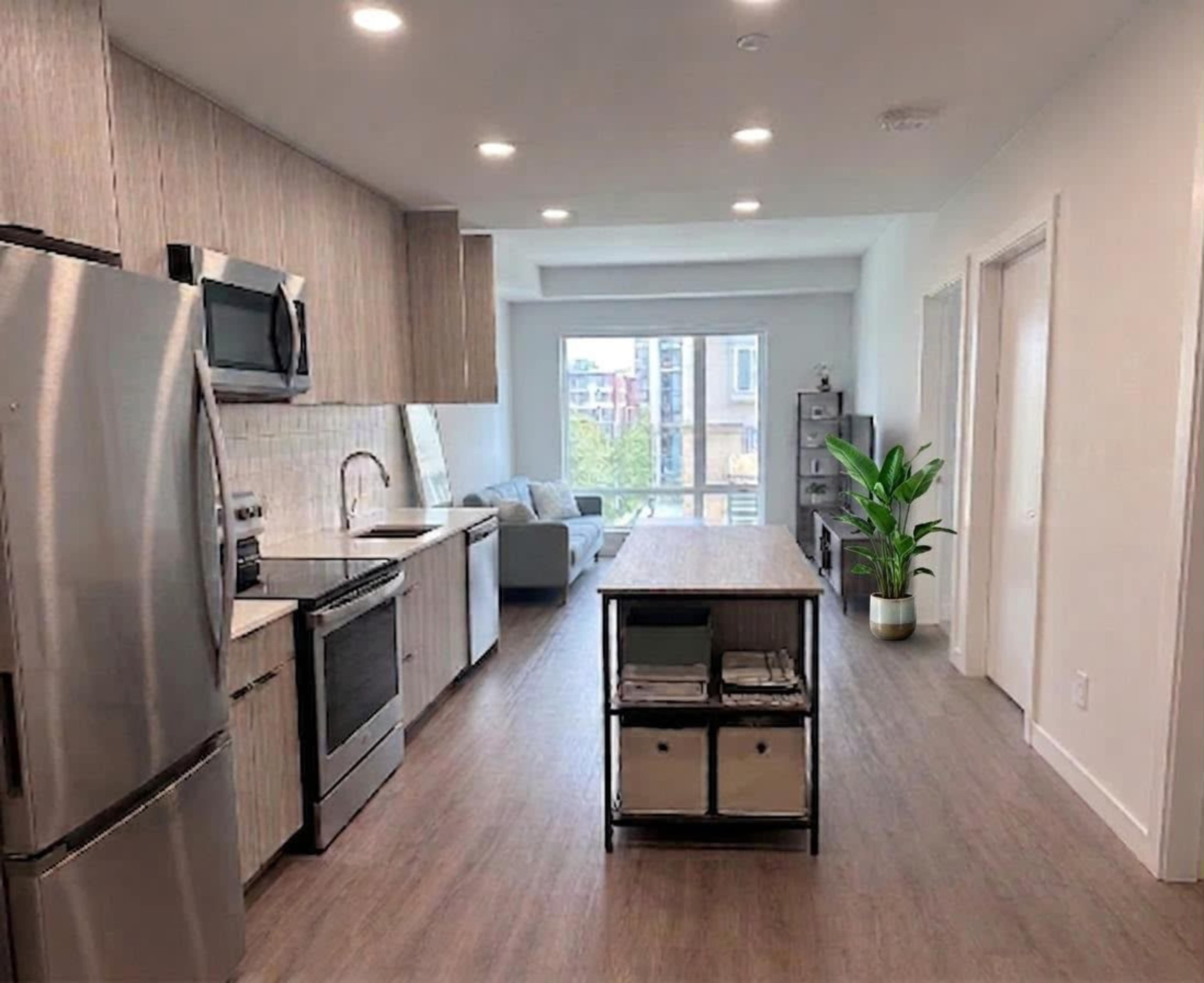 The image shows a modern kitchen with sleek cabinetry, stainless steel appliances, and an island that separates it from a living area with a sofa and a large window.