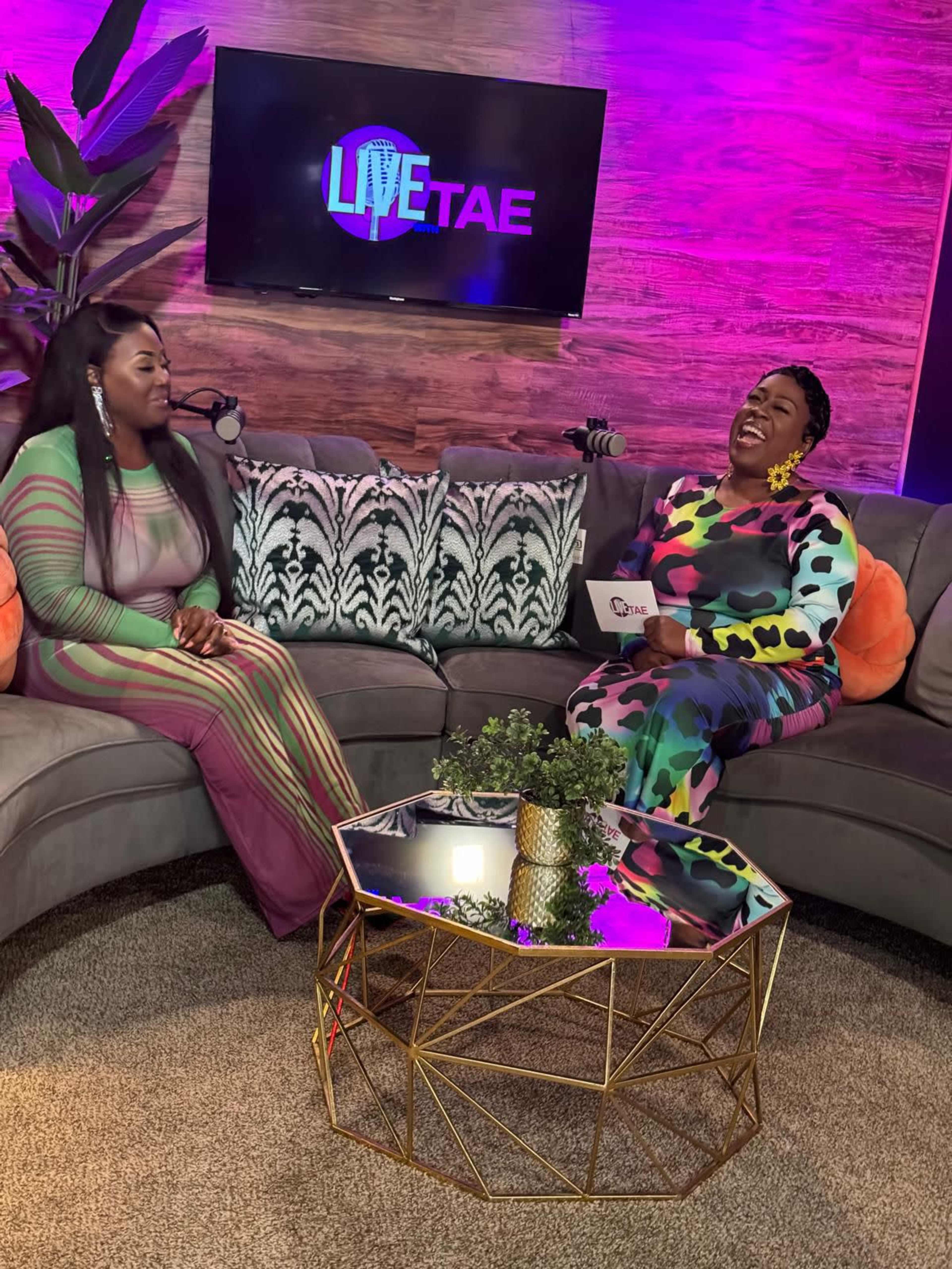 Two women are sitting on a stylish gray sofa, engaging in conversation while a colorful backdrop features the logo "LIVETAE."