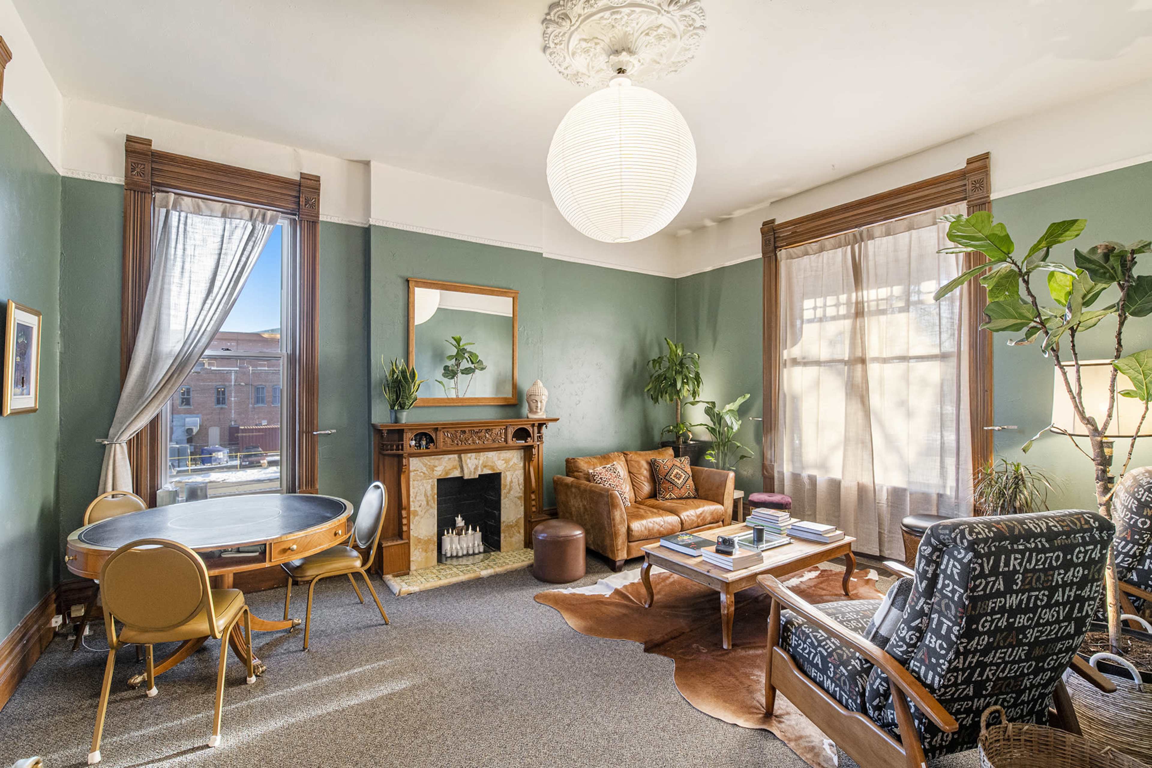 The room features a round table, a leather sofa, and an armchair, with large windows allowing light to illuminate the green walls and a decorative fireplace.
