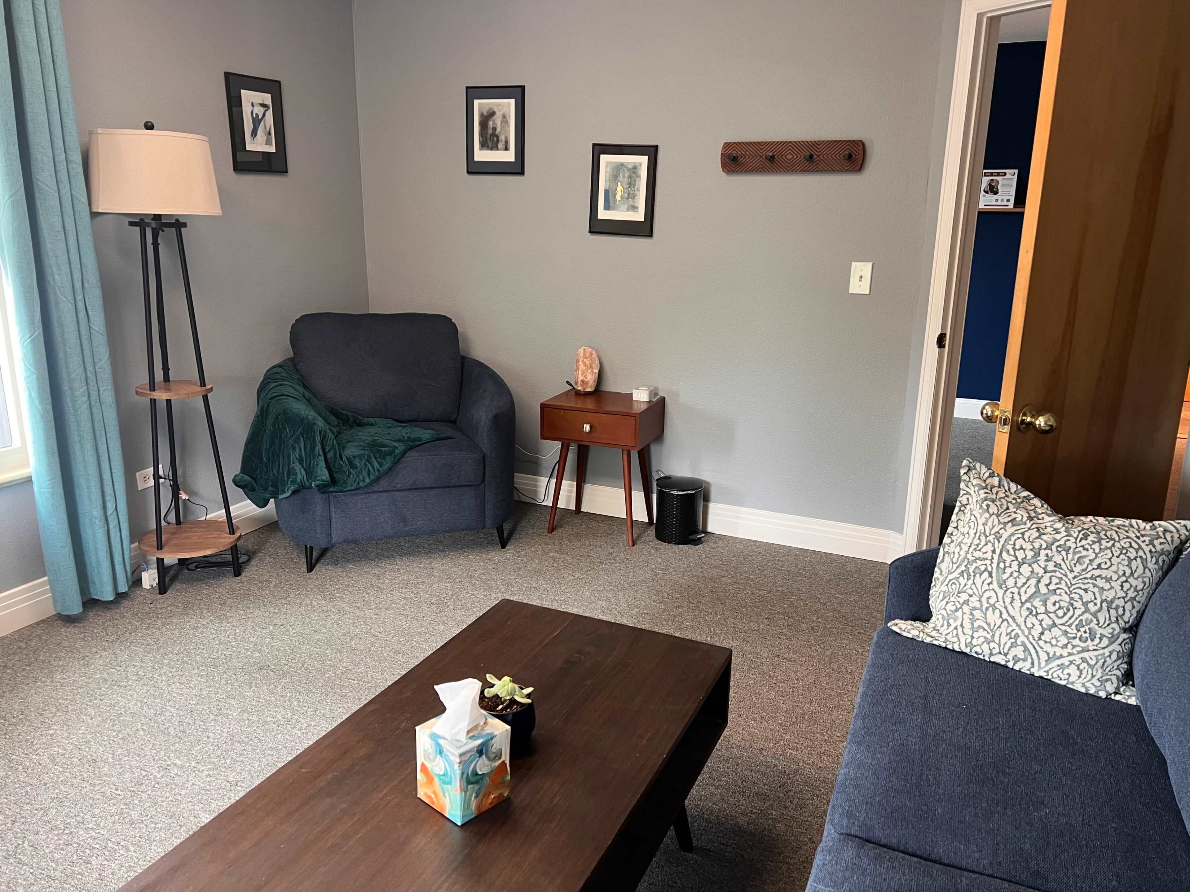 The image shows a cozy living room with a blue armchair, a gray sofa adorned with a patterned pillow, a wooden coffee table, and a lamp beside a window.