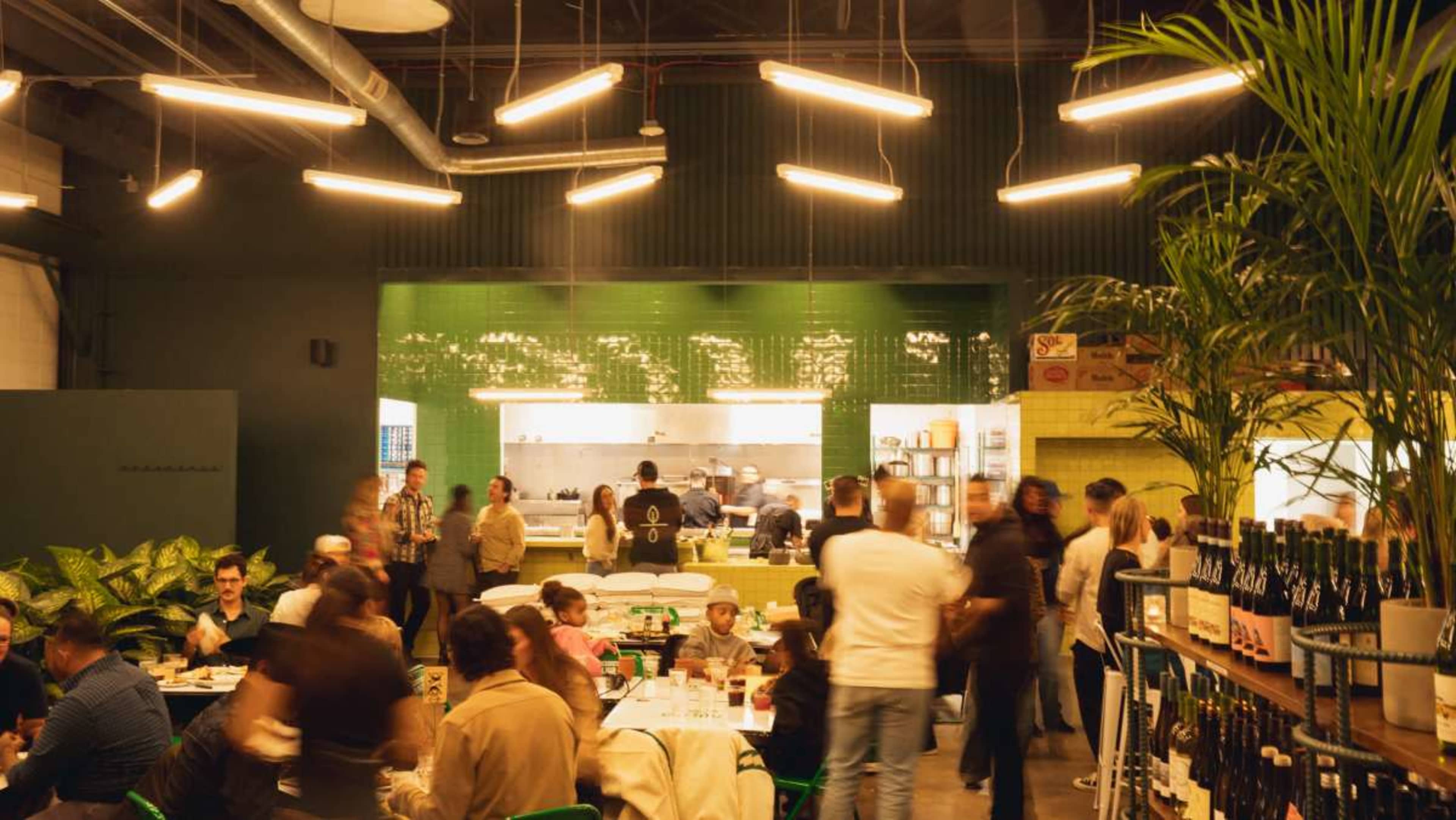 A busy restaurant scene shows patrons dining and interacting under modern lighting, with a visible kitchen in the background.