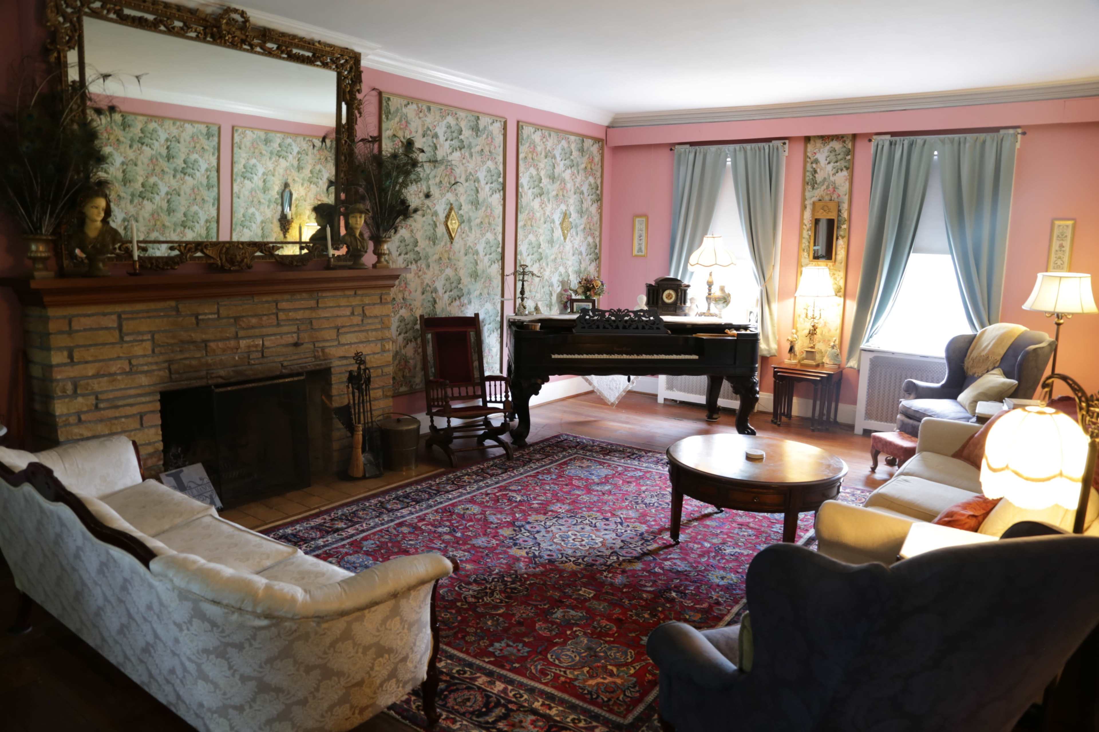 A vintage living room with floral wallpaper, a stone fireplace, and a grand piano, surrounded by various seating options and decorative lamps.