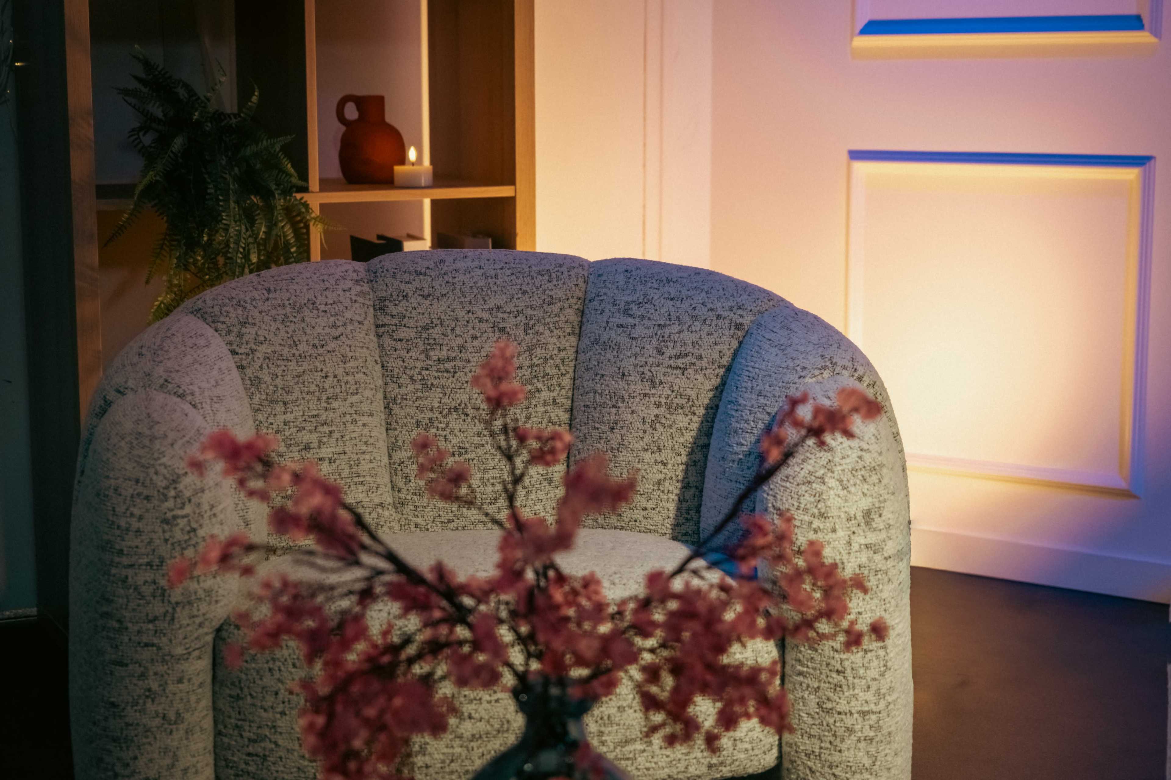 A gray upholstered armchair is positioned near a softly lit shelf, with a decorative vase of pink flowers in the foreground.