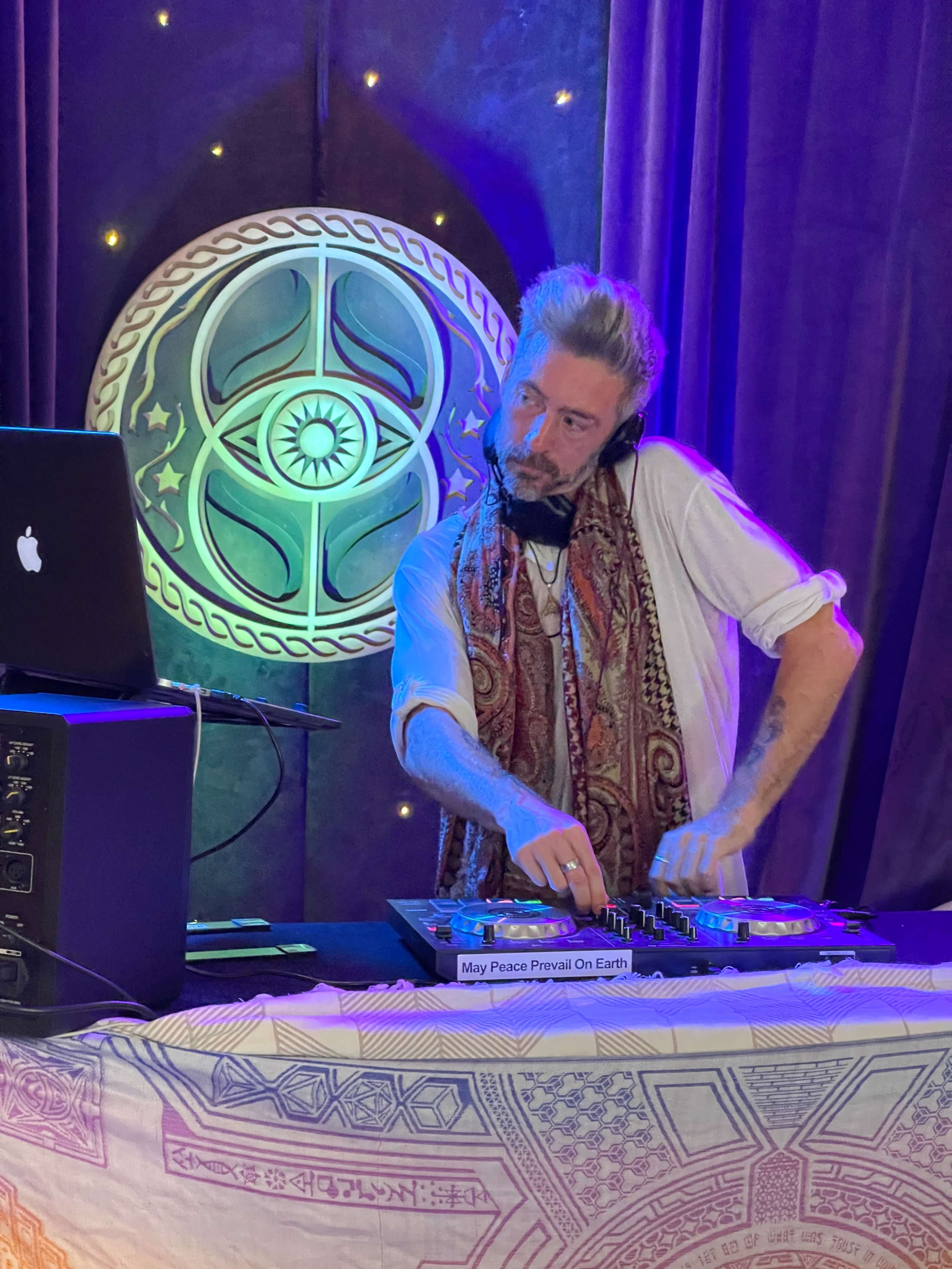 A DJ stands behind a table with turntables and a laptop, mixing music under colorful lights and a decorative backdrop.
