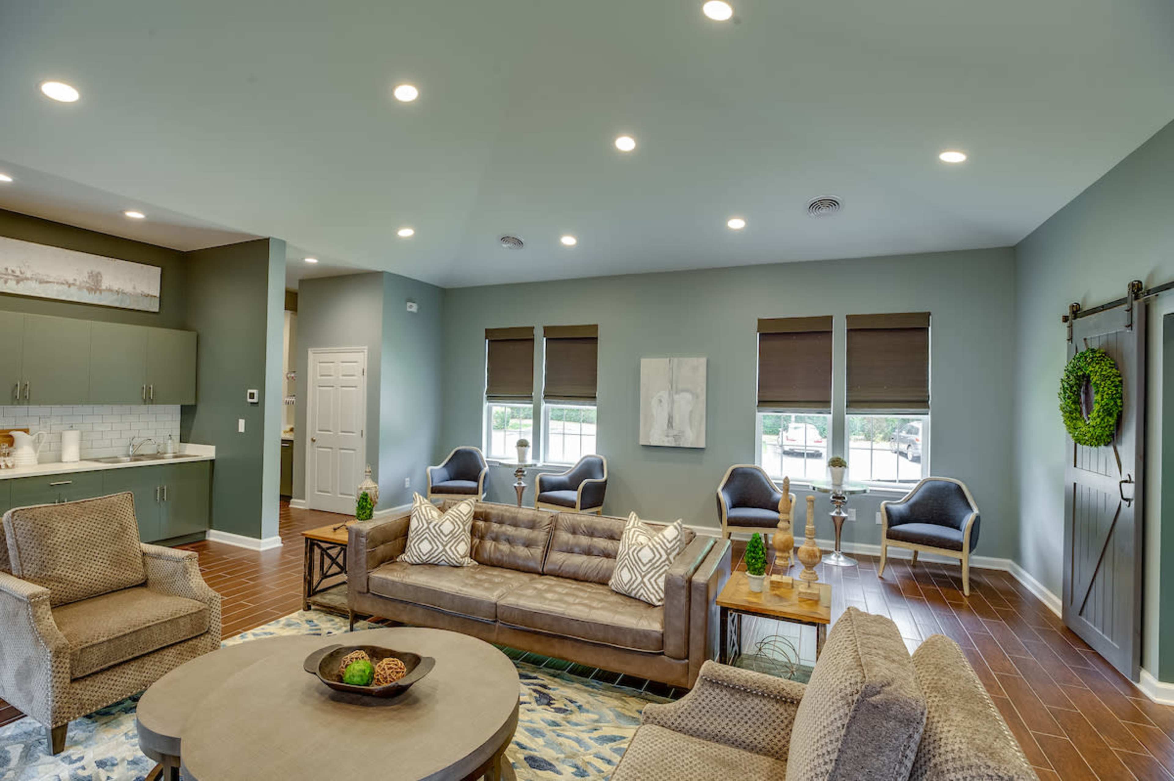 A modern living room with a sectional sofa, a round coffee table, and several chairs, all arranged around a patterned area rug, with a kitchenette visible in the background.