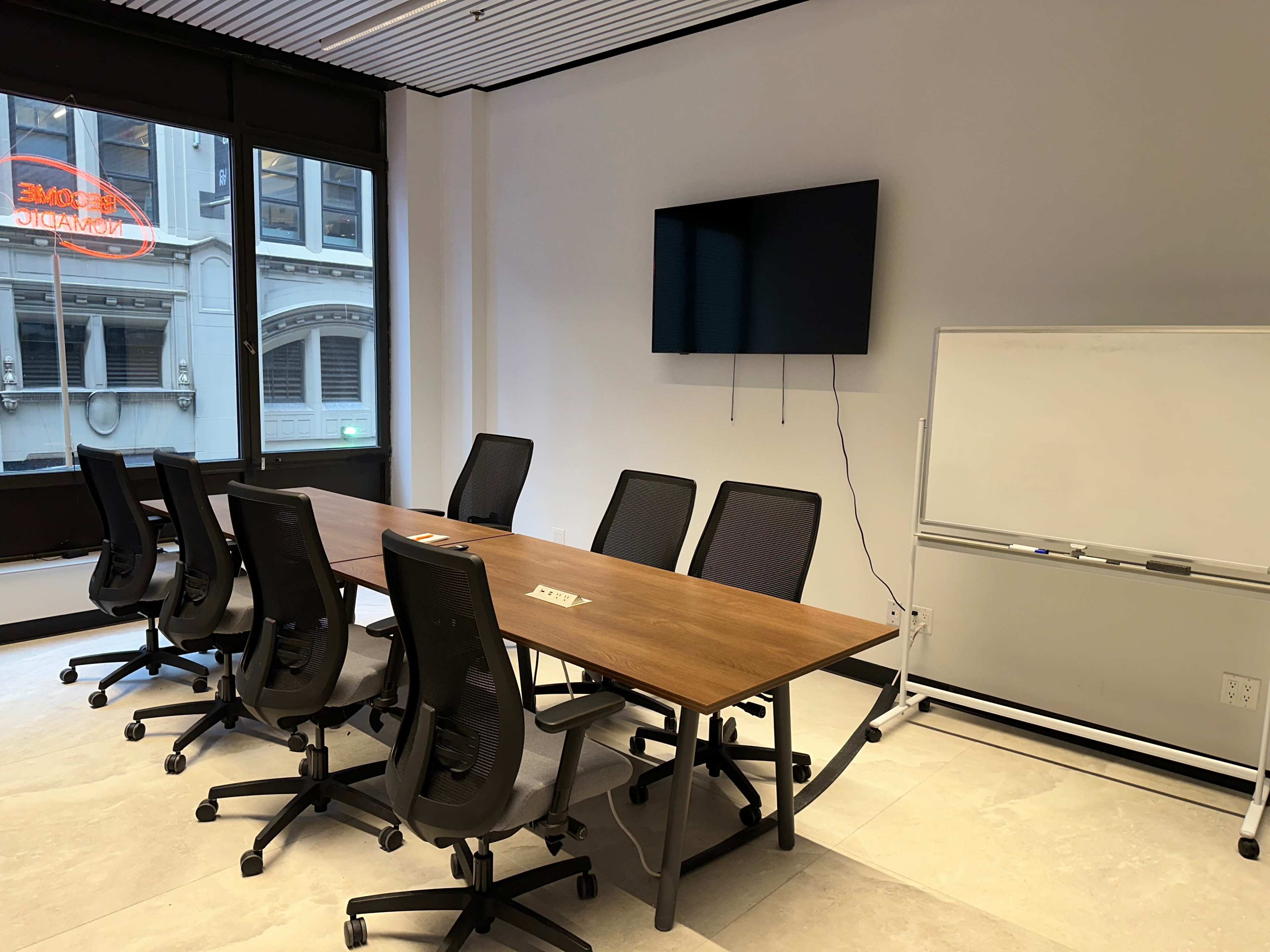 A conference room features a long wooden table surrounded by black ergonomic chairs, a large screen mounted on the wall, and a whiteboard nearby.