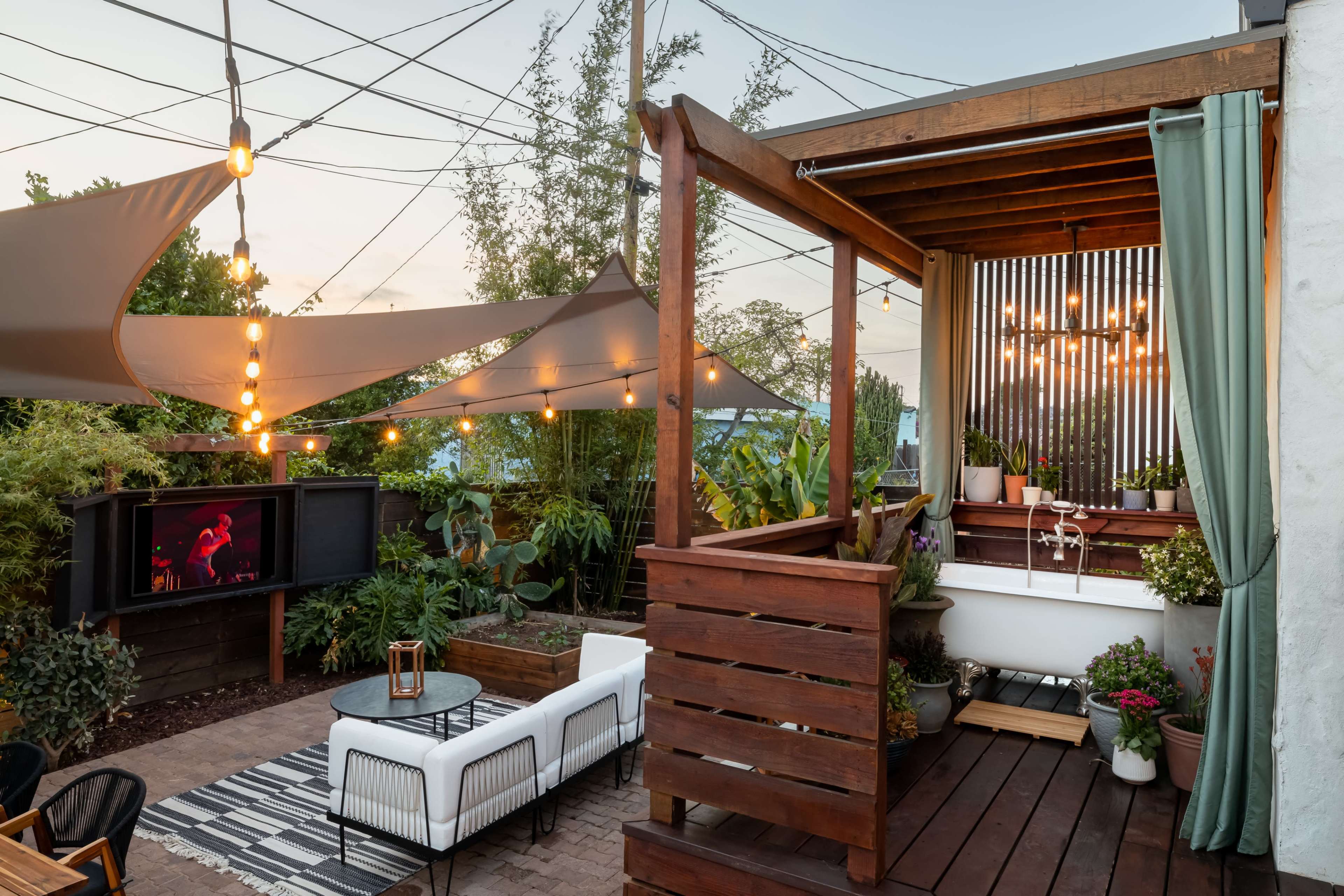 The image shows a backyard with a wooden deck, outdoor seating, and a large television, surrounded by plants and illuminated by string lights.