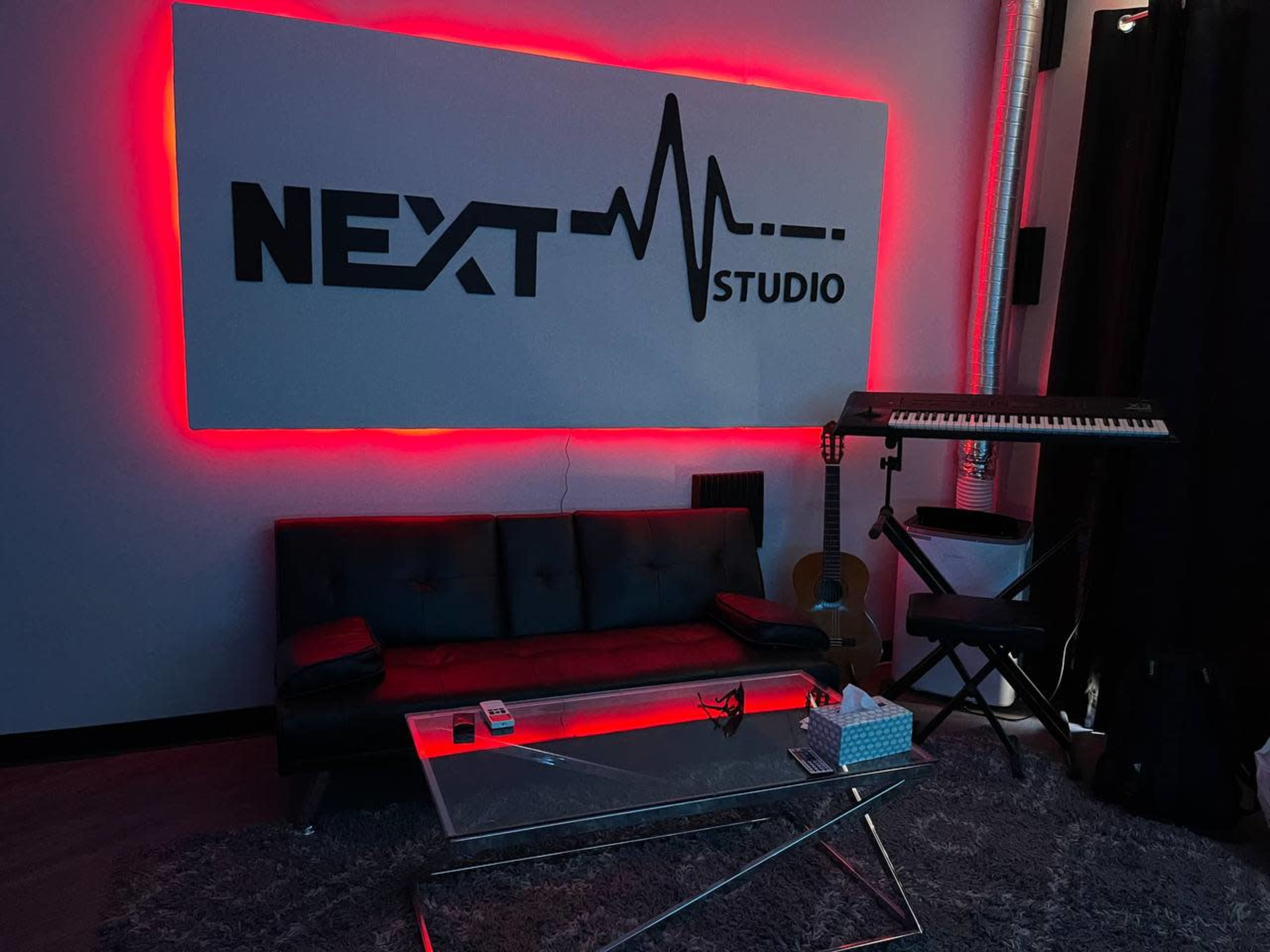 A modern studio space featuring a black sofa, a glass coffee table, a keyboard on a stand, and red accent lighting around the wall.