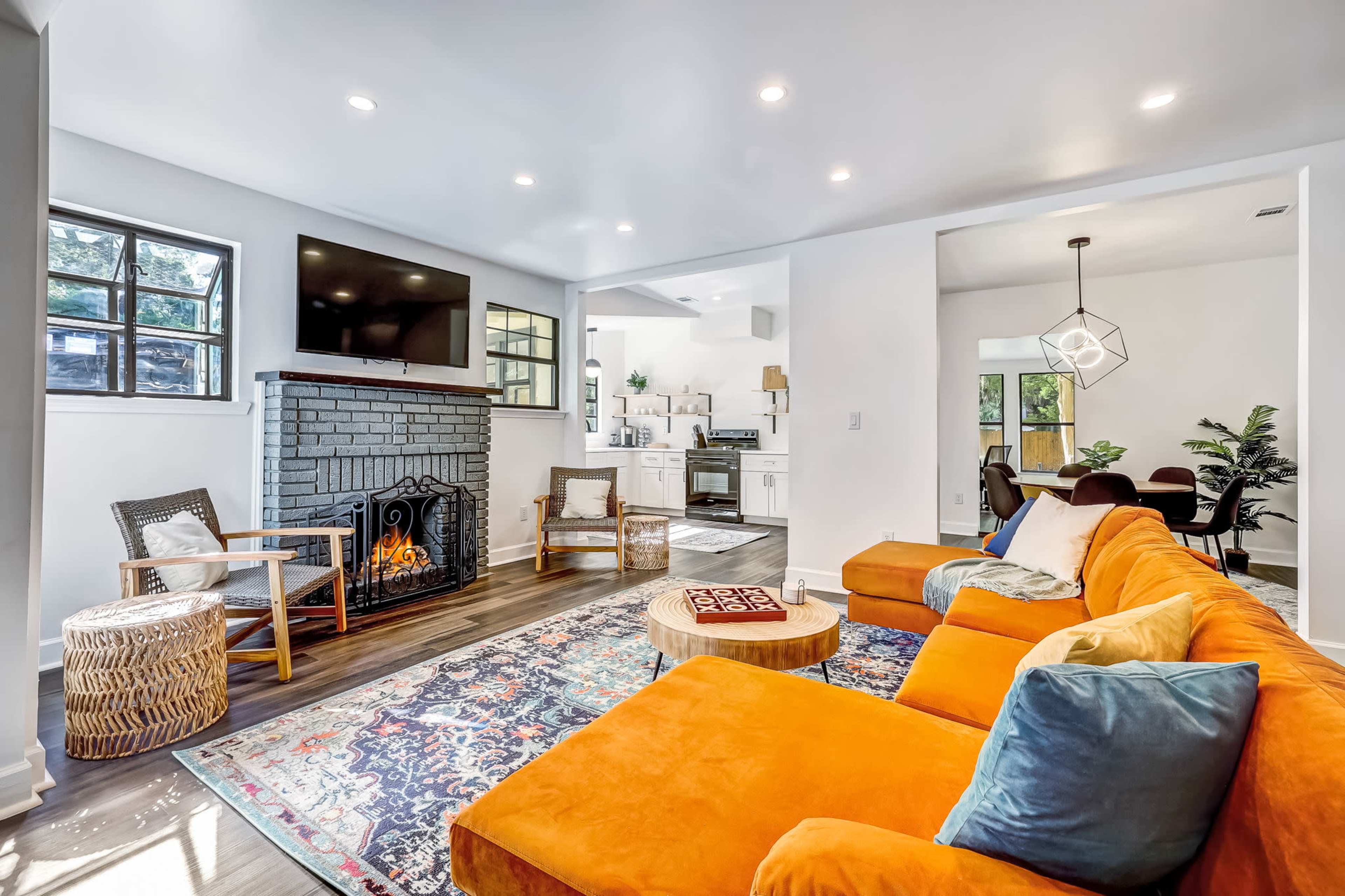 A living room features an orange sectional sofa, a round coffee table, and a fireplace, with a television mounted above and a view into a kitchen area.