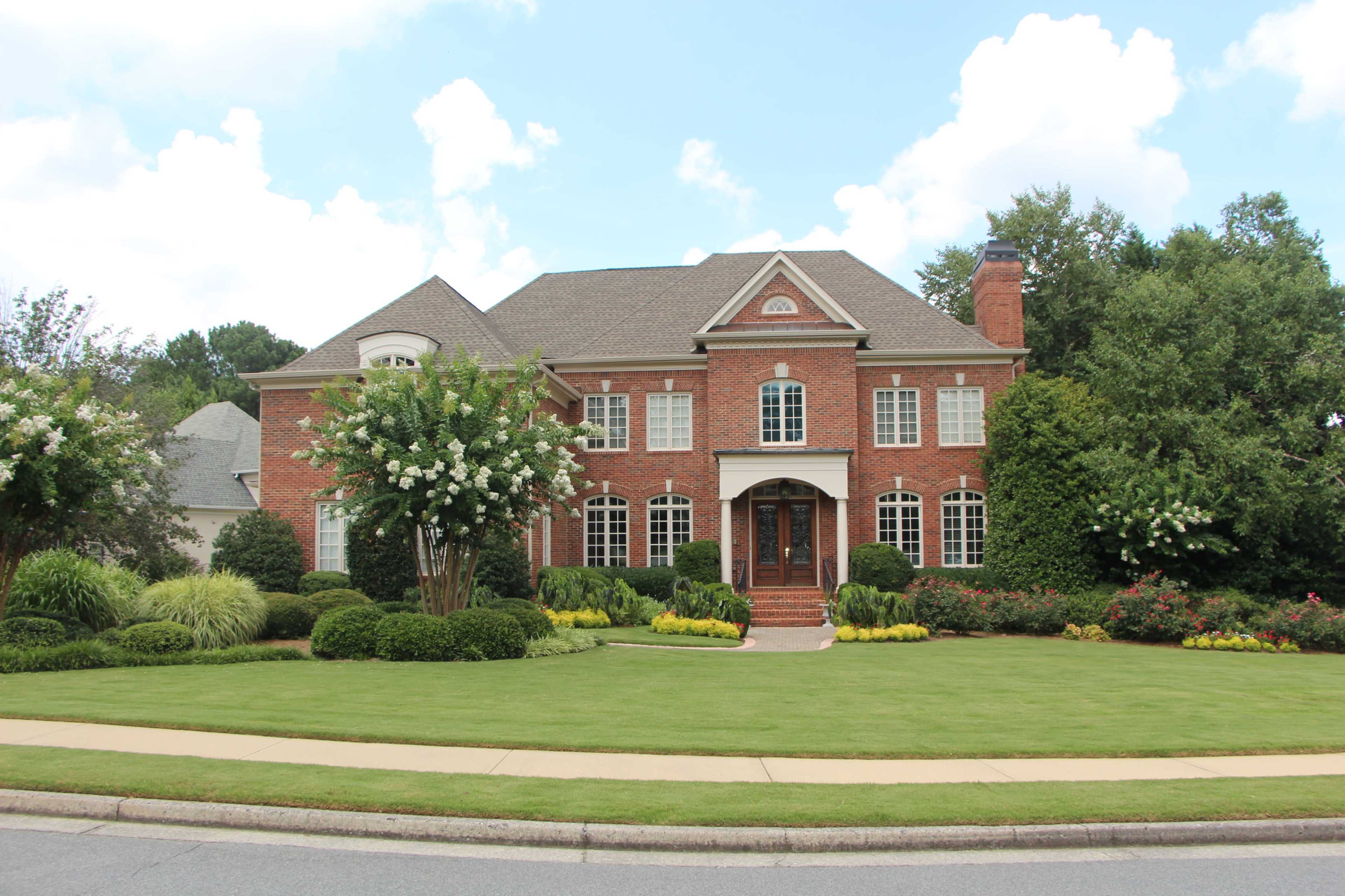 The image shows a large, red-brick two-story house surrounded by neatly trimmed hedges and a well-maintained lawn.