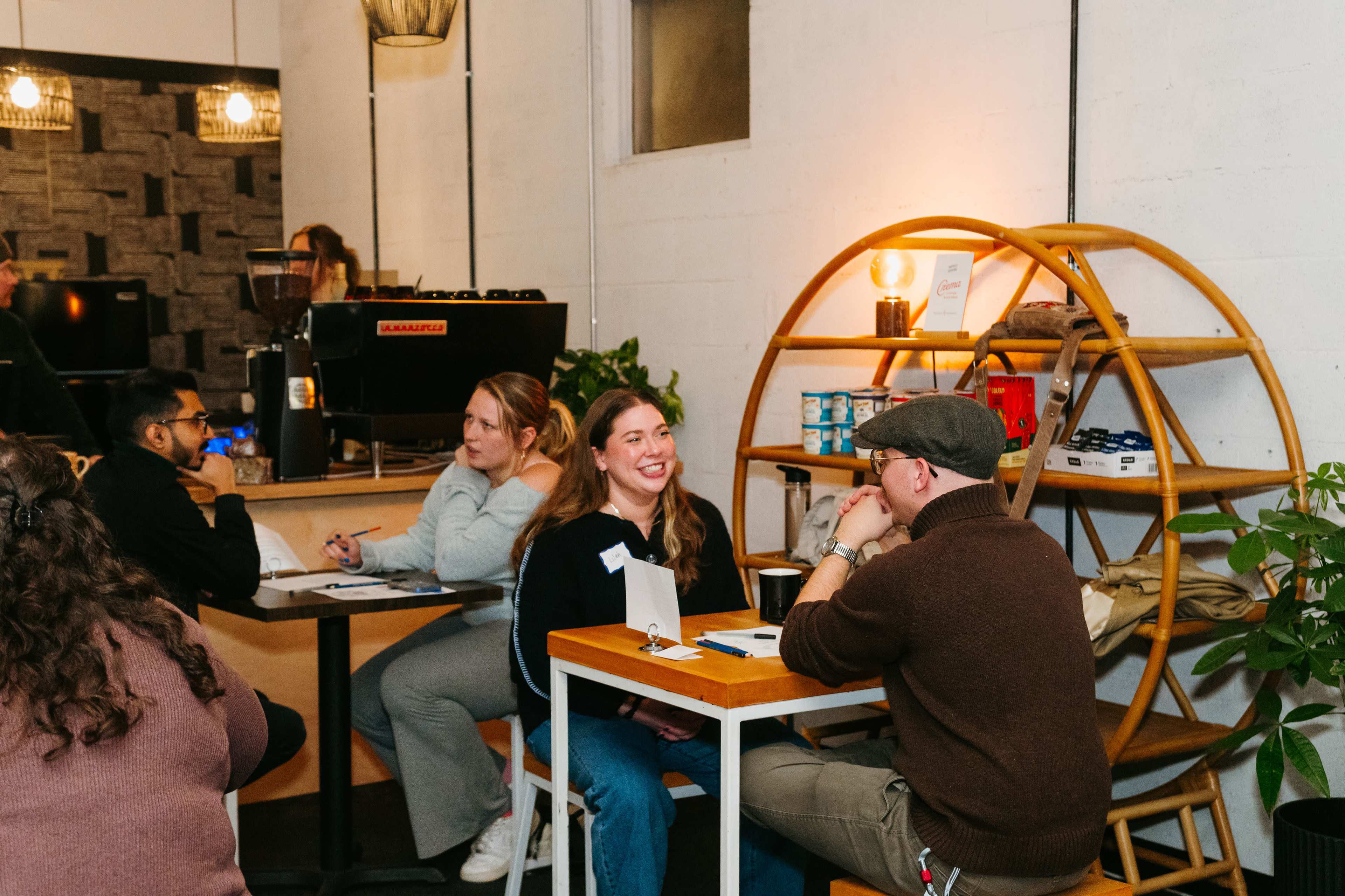 A group of people sits at tables in a cozy cafe, engaged in conversation, with a bookshelf and plants in the background.