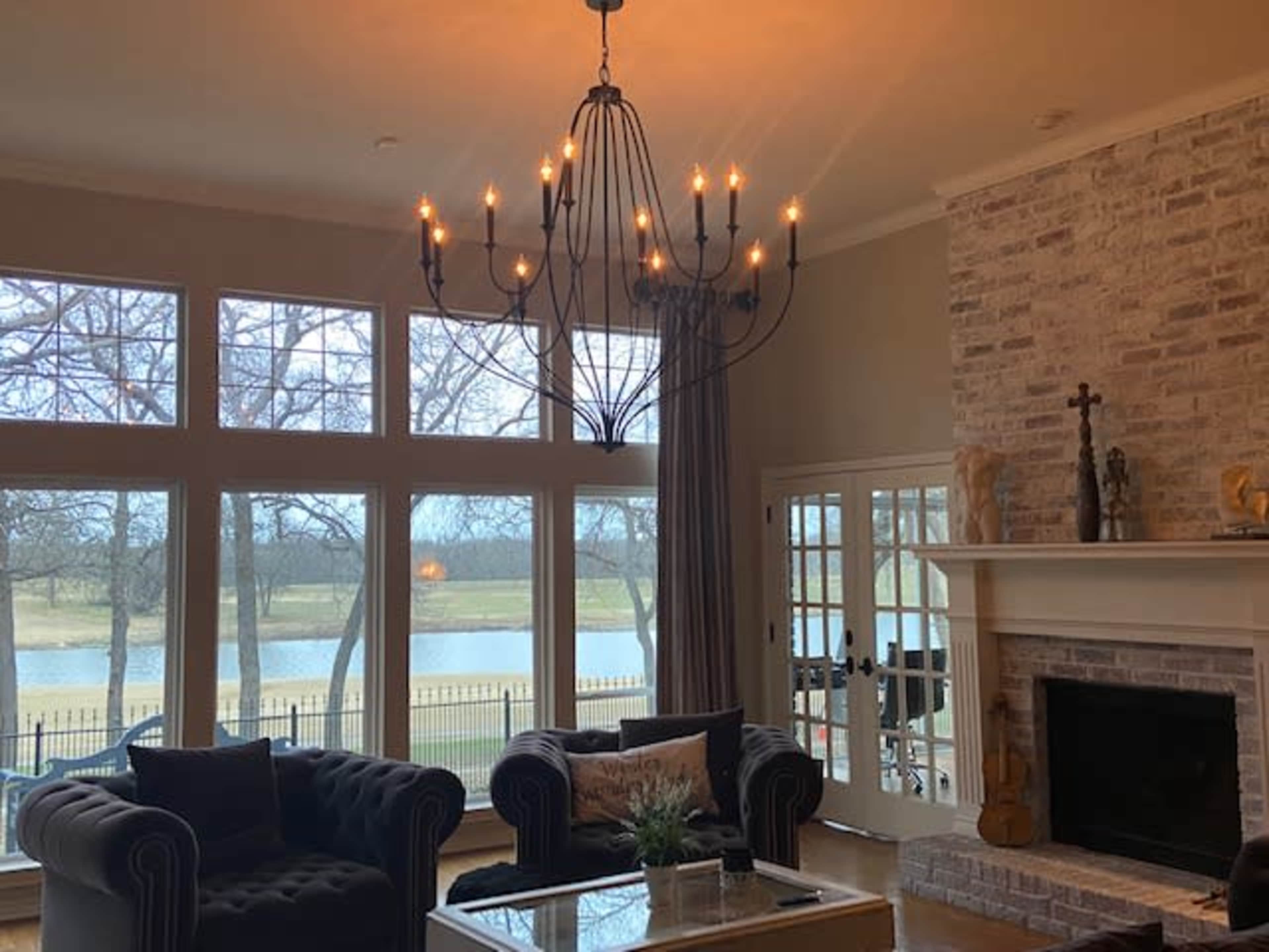 A living room features large windows overlooking a lake, with a chandelier hanging above a coffee table and two tufted sofas.