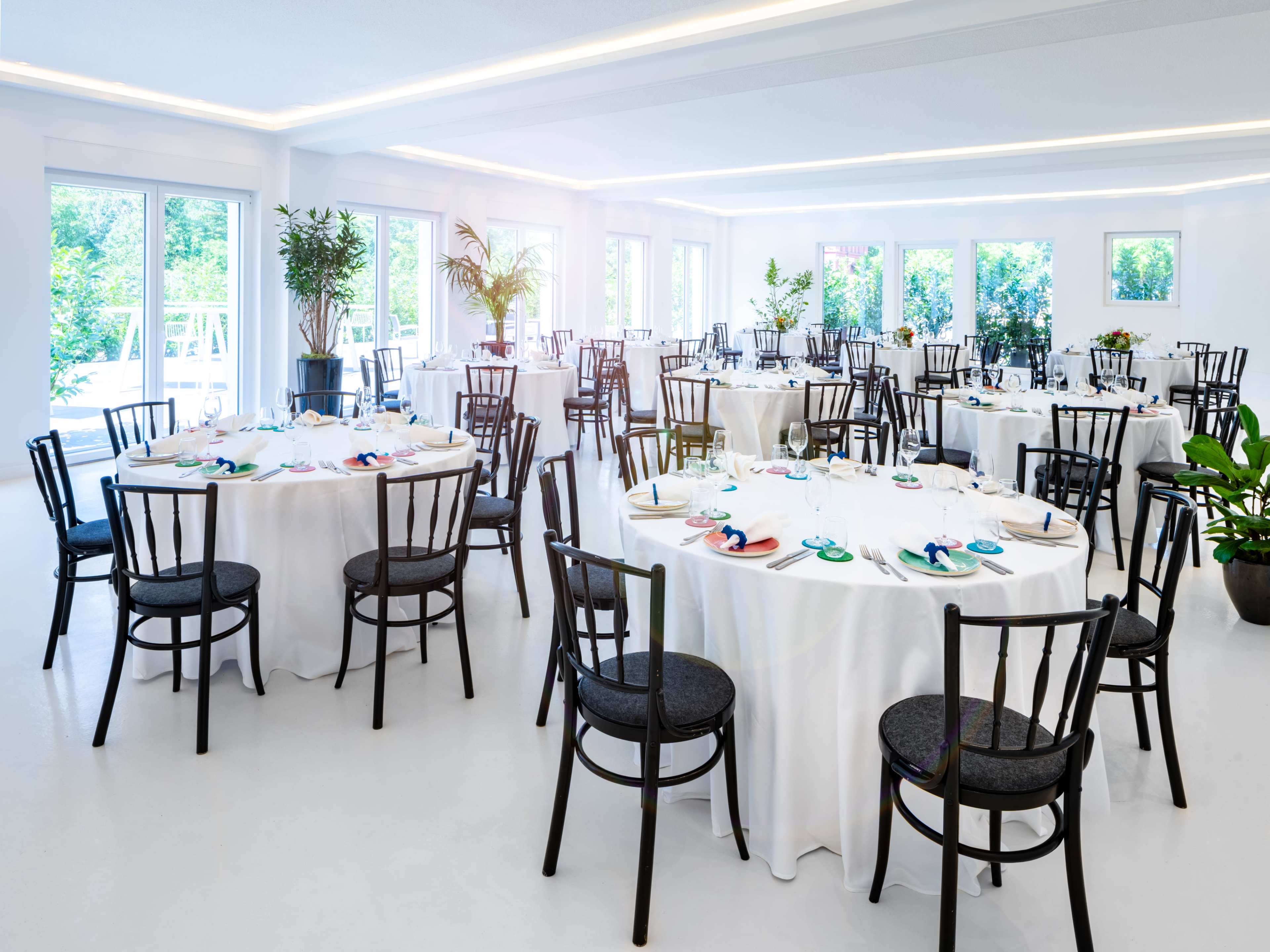A spacious dining room features several round tables set with white tablecloths and black chairs, surrounded by greenery and large windows.