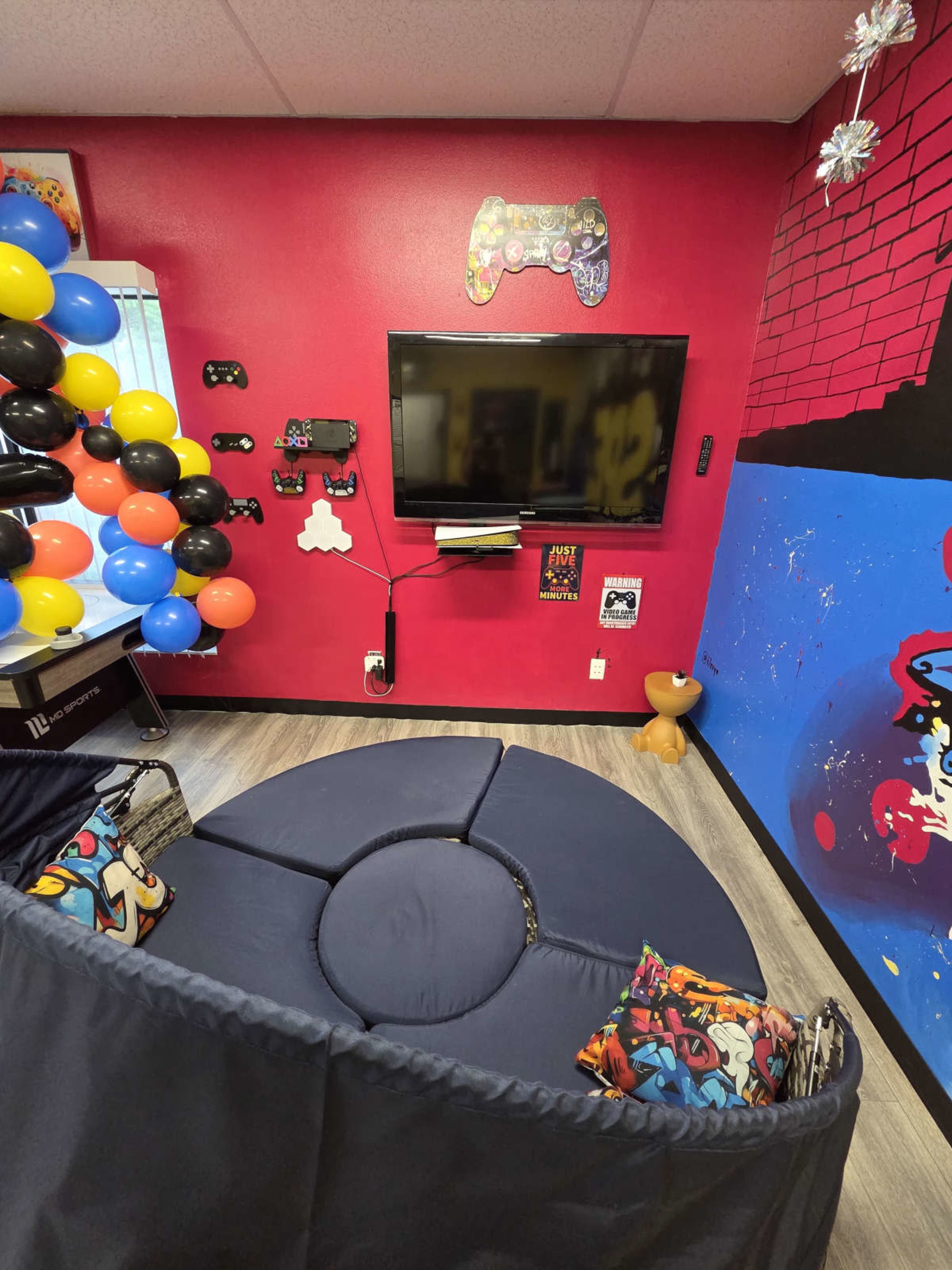 The image features a modern gaming lounge with a circular navy-blue seating area, a wall-mounted TV, and colorful decorations, including balloons and artwork.