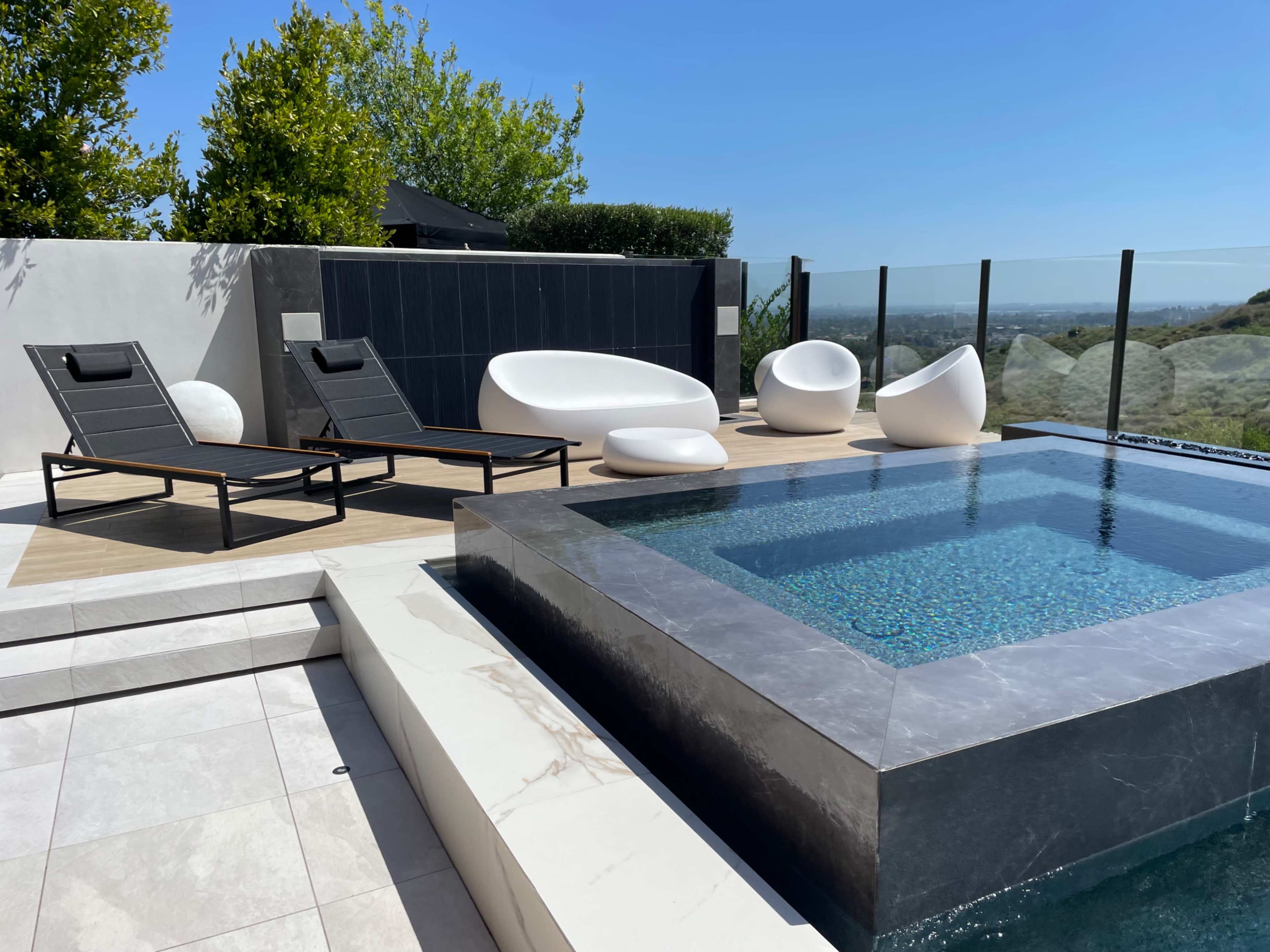 The image shows a modern pool area featuring a rectangular swimming pool, lounge chairs, and abstract seating against a backdrop of clear blue skies and greenery.