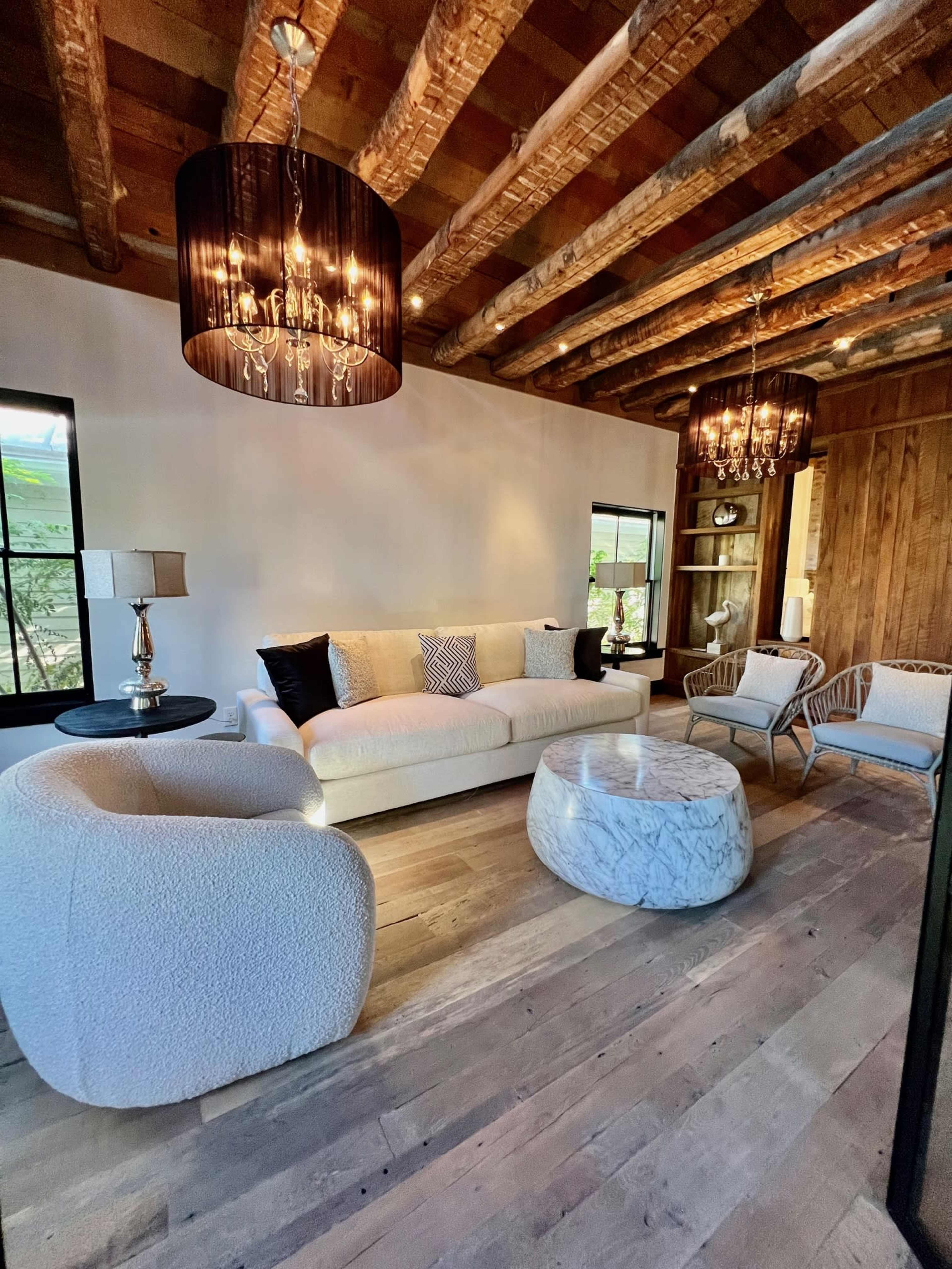 A stylish living room featuring a large, light-colored sofa, a round marble coffee table, and two modern chairs under decorative chandeliers with wooden beams overhead.