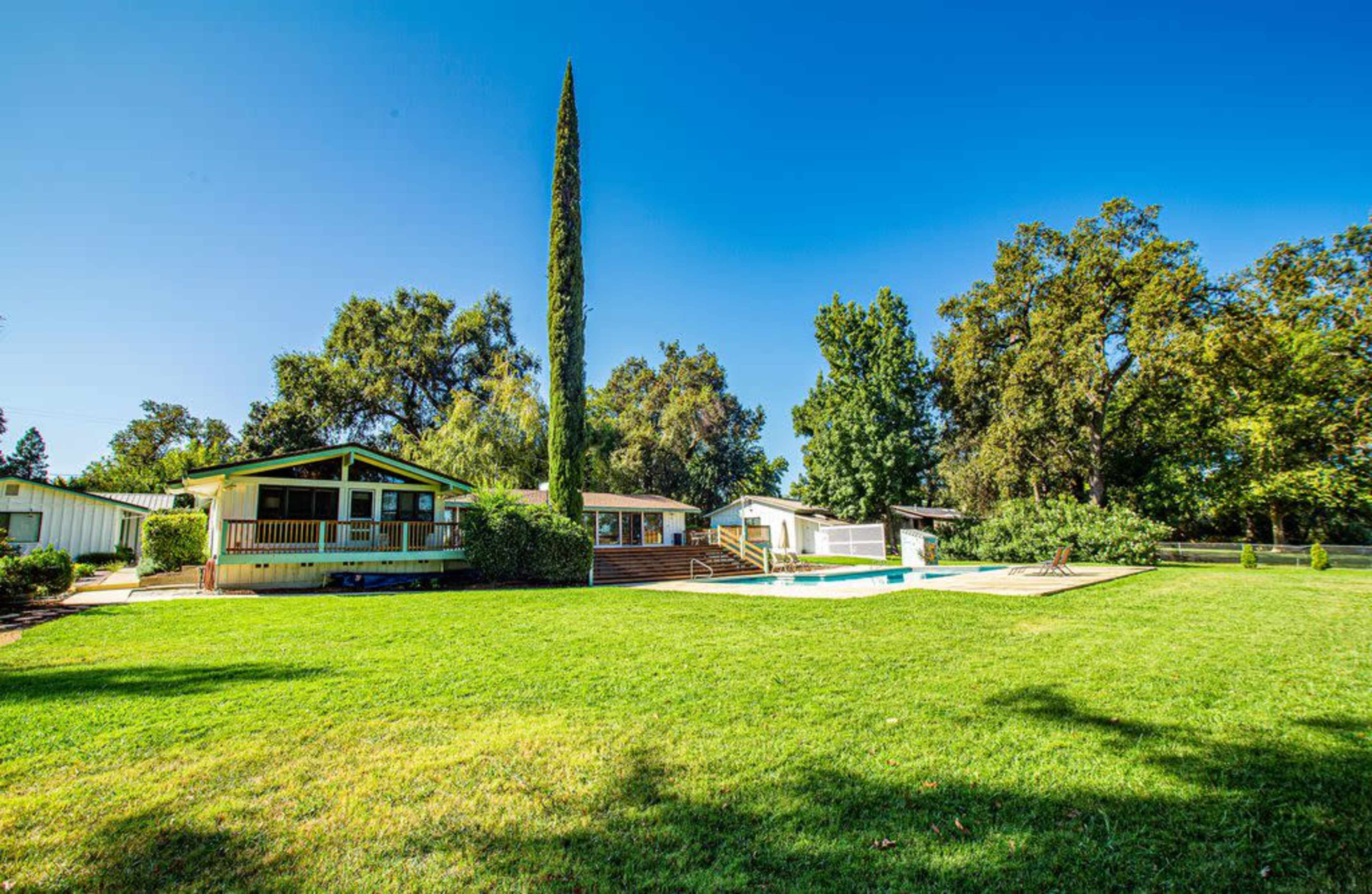 A lush green lawn features a swimming pool and several houses surrounded by trees in a sunny outdoor setting.