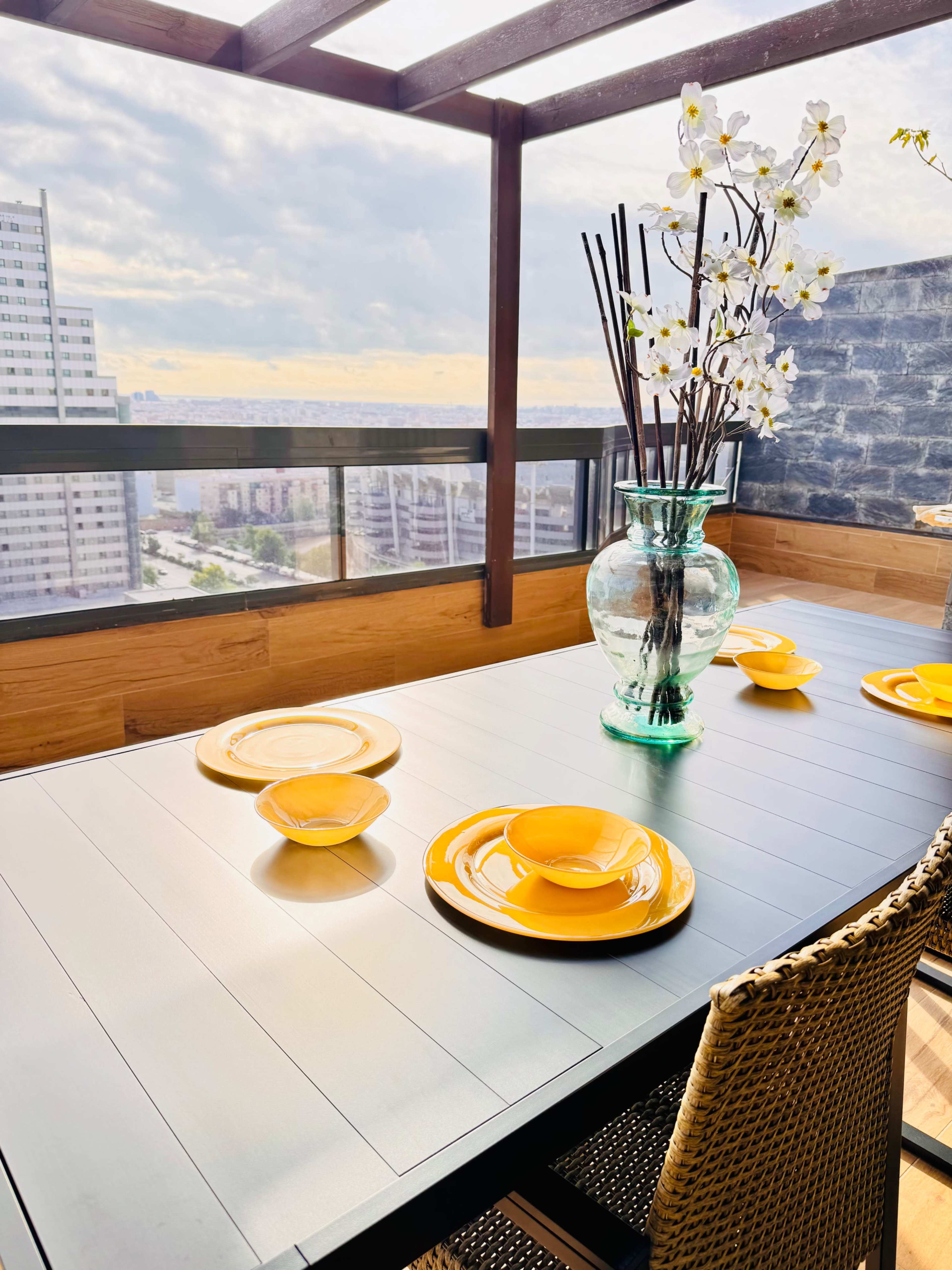 A table is set with yellow plates and bowls, adorned with a large vase of decorative flowers, and overlooks a cityscape from a balcony.