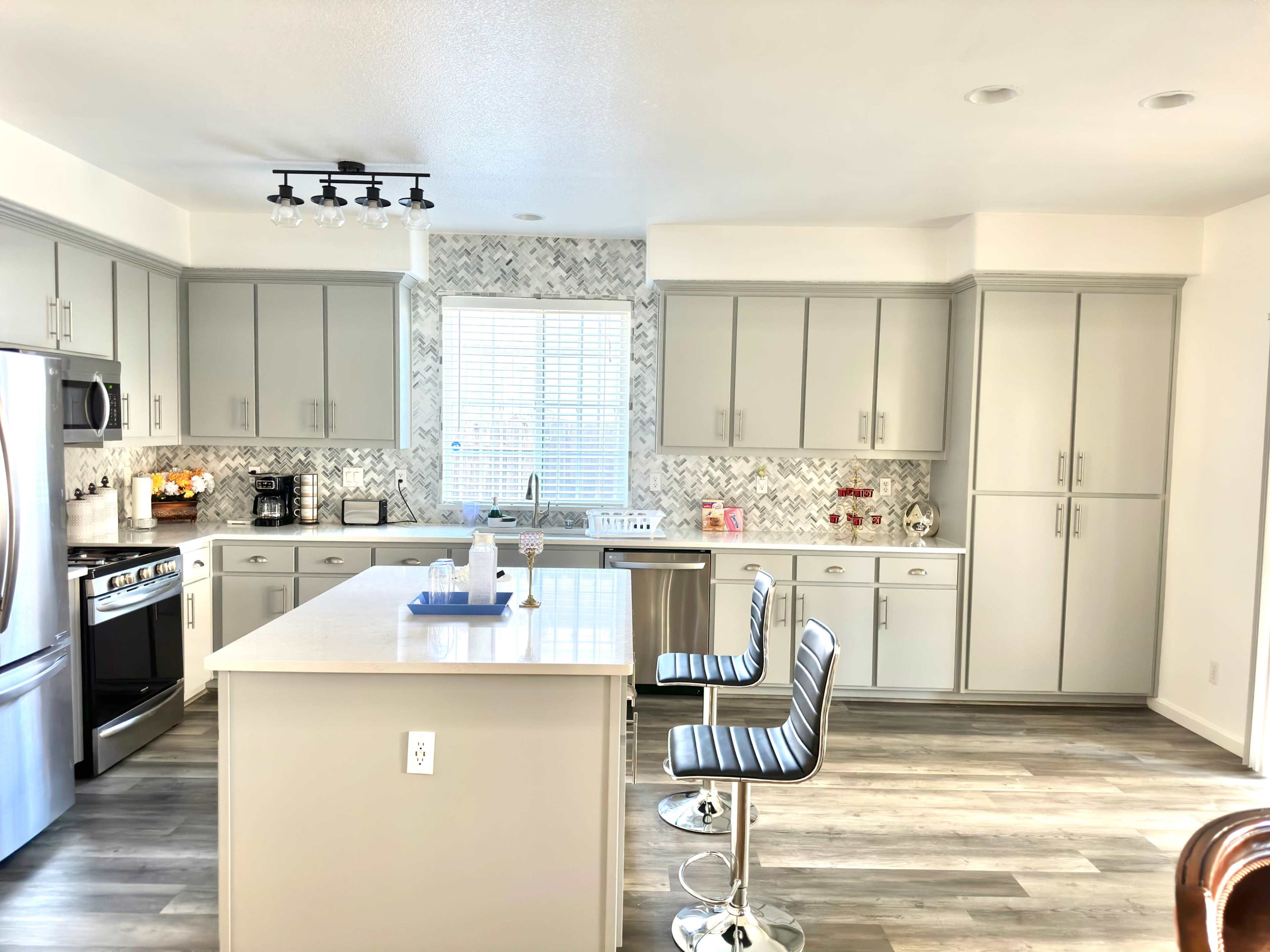 A modern kitchen features gray cabinets, a central island with bar stools, and a window with a view of the outdoors.