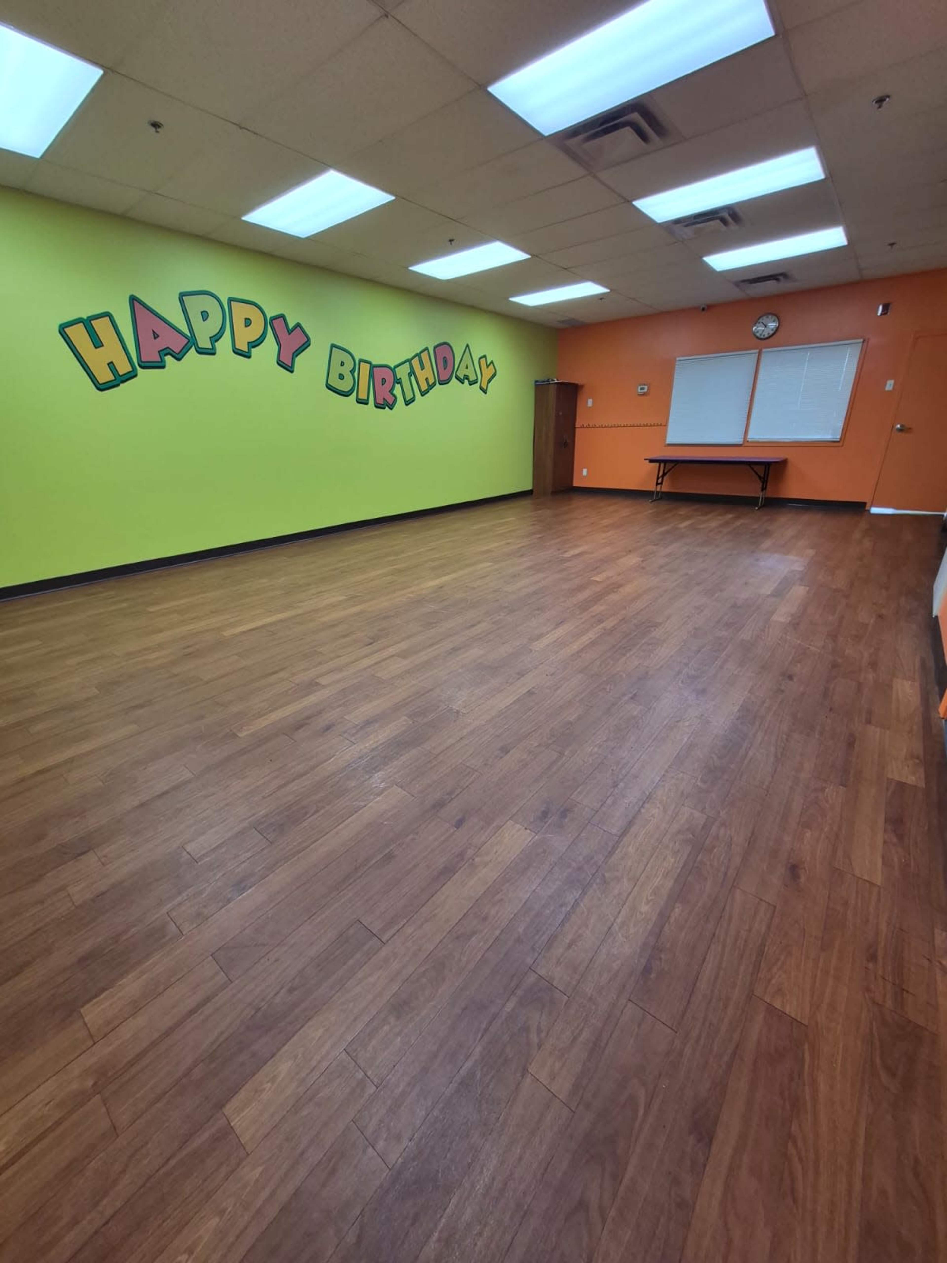 The image shows a large, brightly colored room with green and orange walls, featuring the words "HAPPY BIRTHDAY" painted on one wall and wooden flooring.