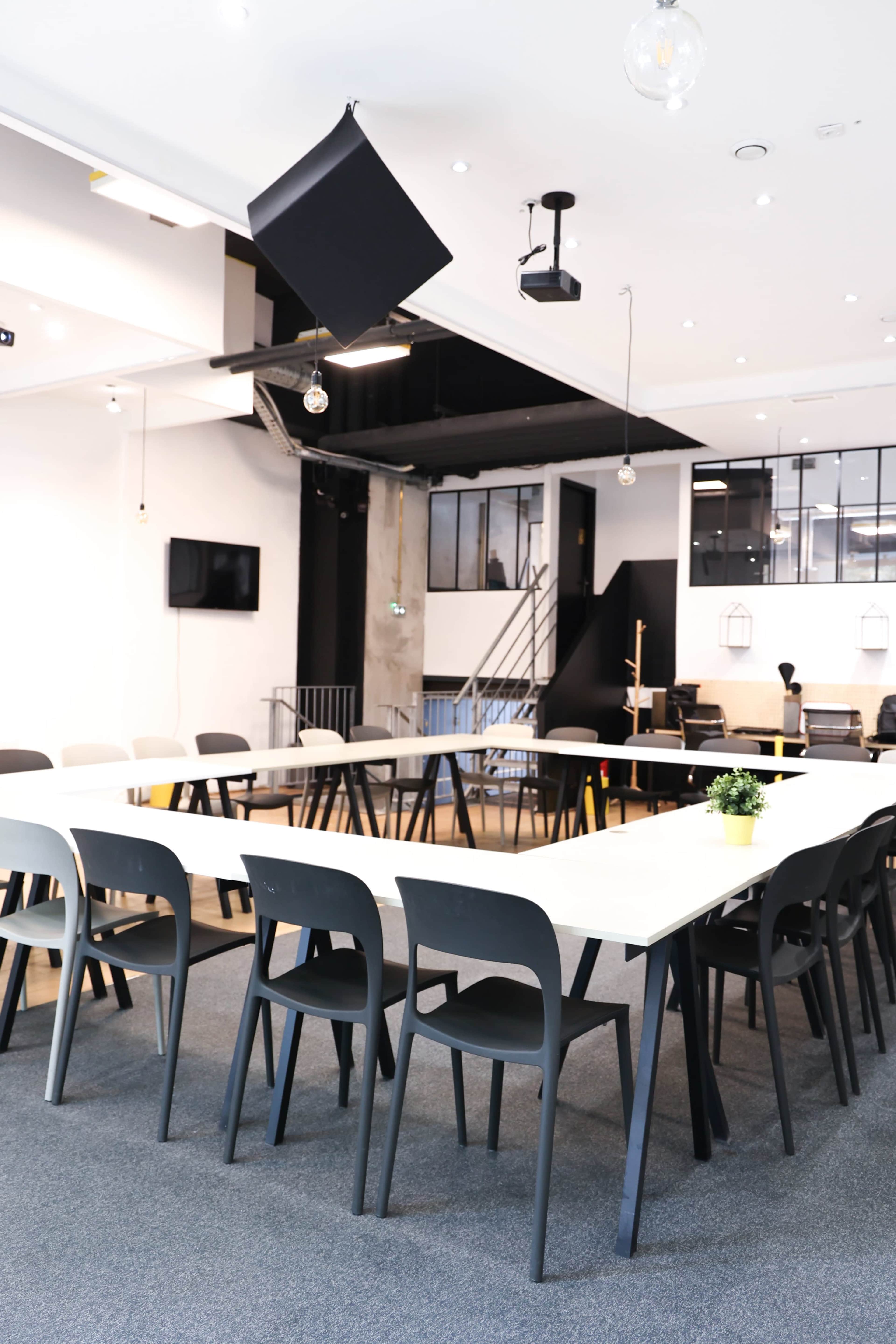 The image depicts a modern conference room with a long table surrounded by black chairs, illuminated by pendant lights and featuring a projector mounted on the ceiling.