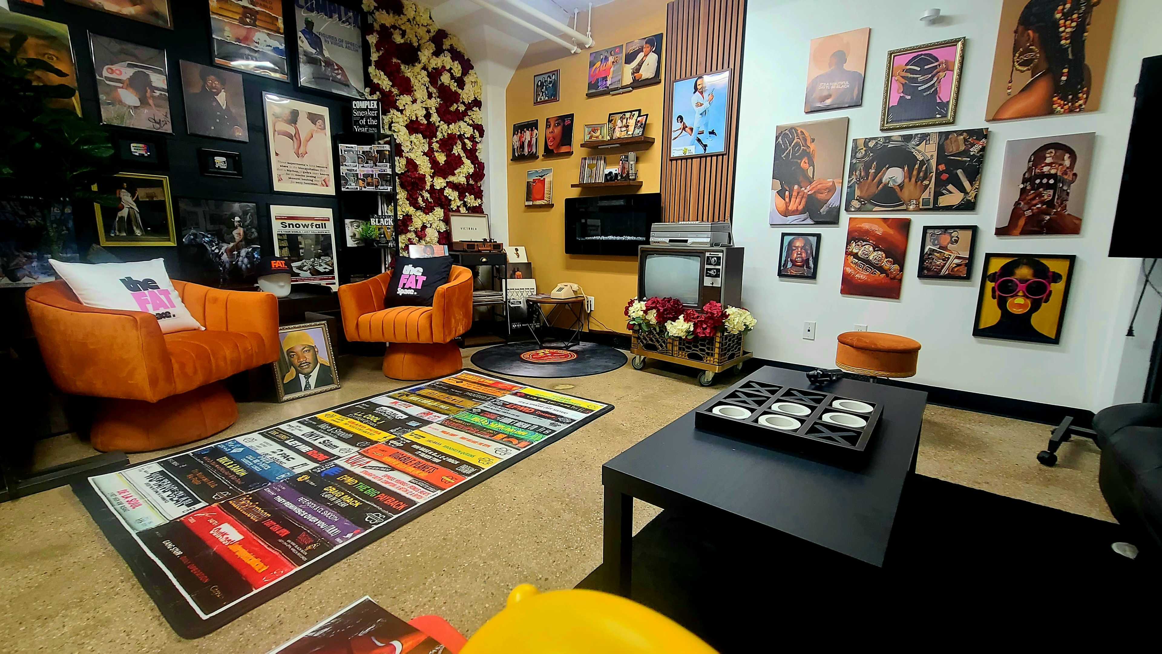 The room features two orange chairs, a black record player on the floor, a wall adorned with framed photographs and art, and a colorful area rug with a grid of album covers.