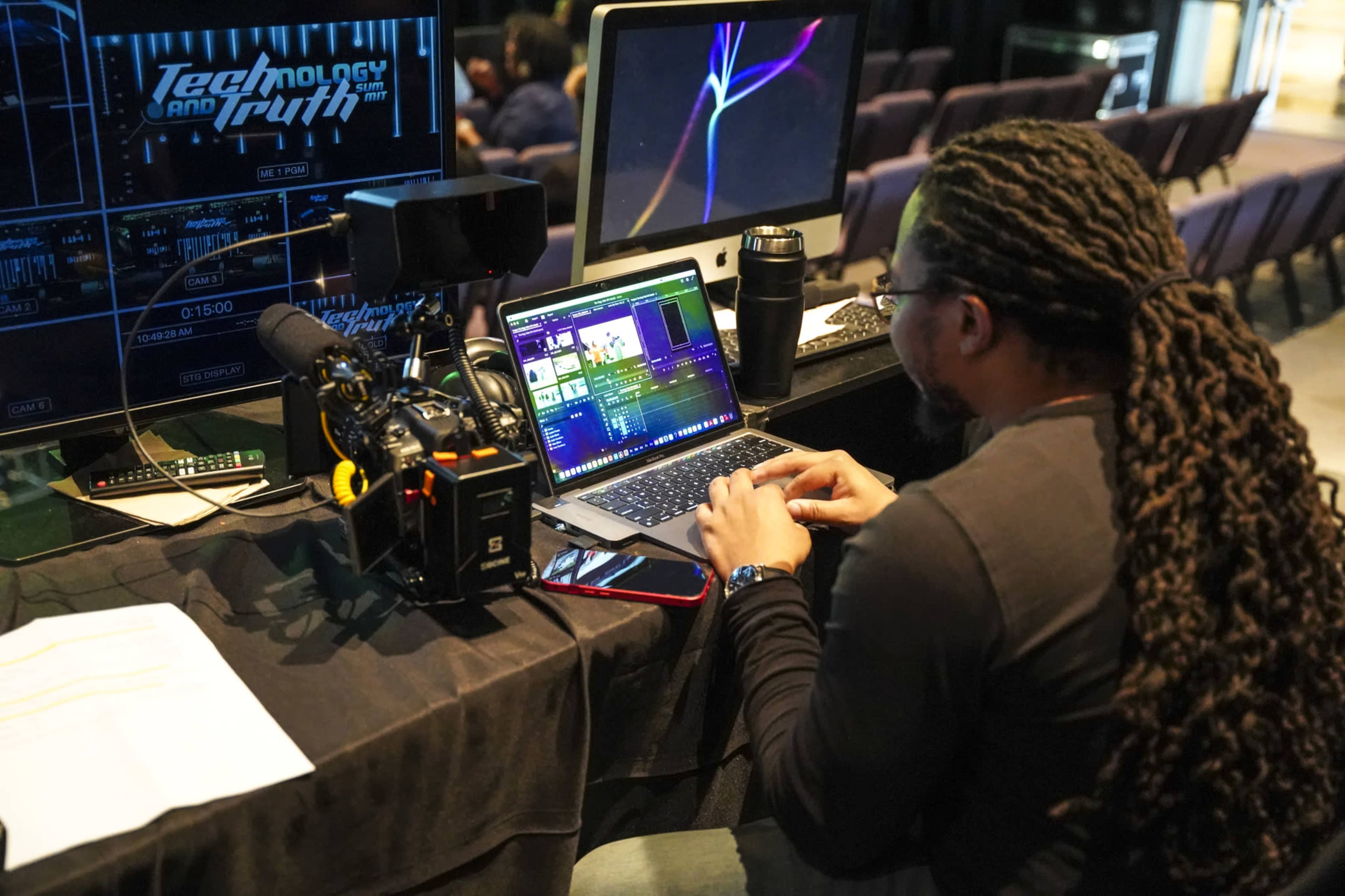 A person with long hair works on video editing software on a laptop while seated at a desk with a camera and monitors displaying graphics.