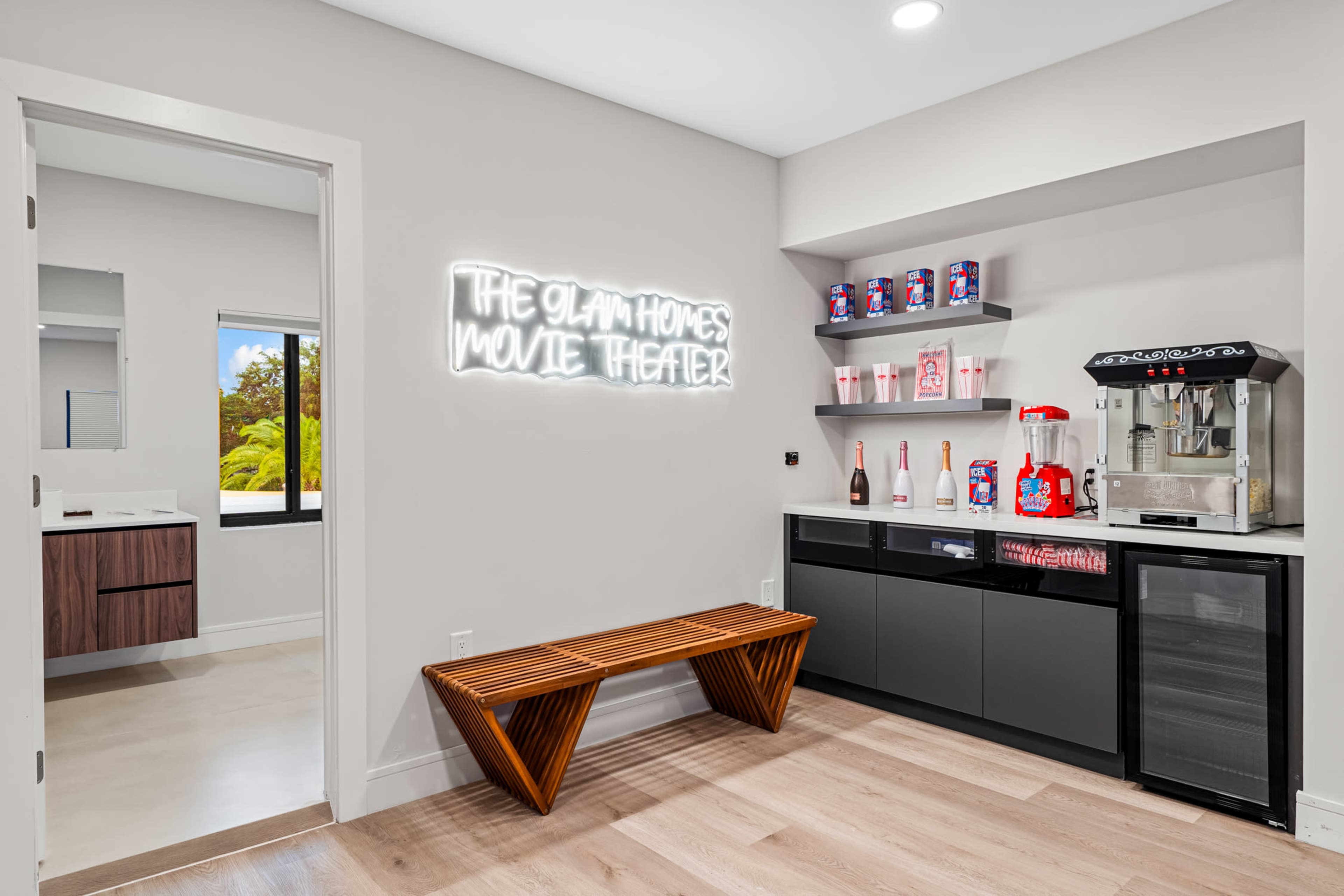 The image shows a modern movie theater room featuring a countertop with a popcorn machine and display shelves, complemented by a neon sign that reads "THE GLAM HOMES MOVIE THEATER."