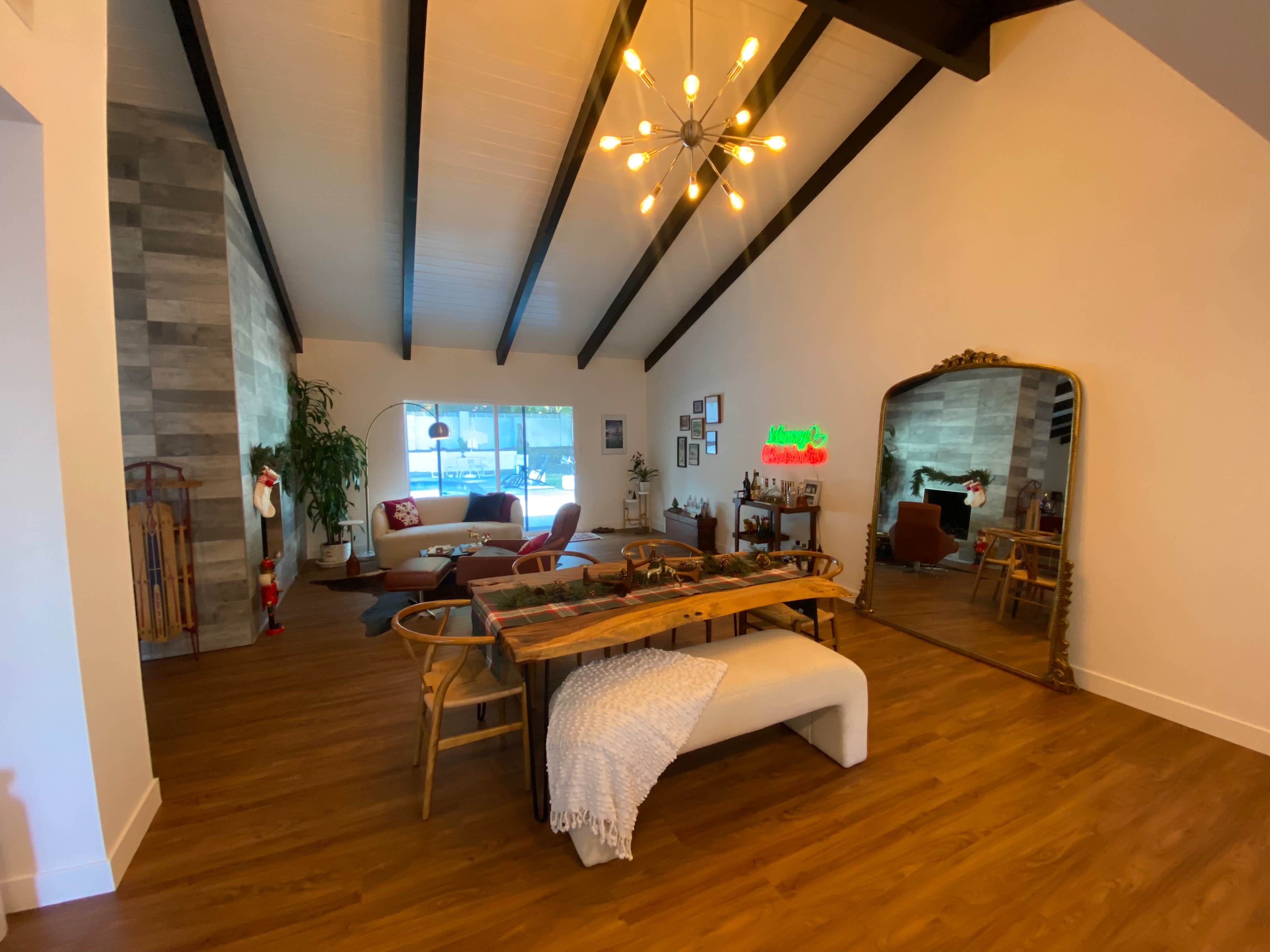 The image shows a modern living space with a high-beamed ceiling, a dining table, a large mirror, and various seating areas.