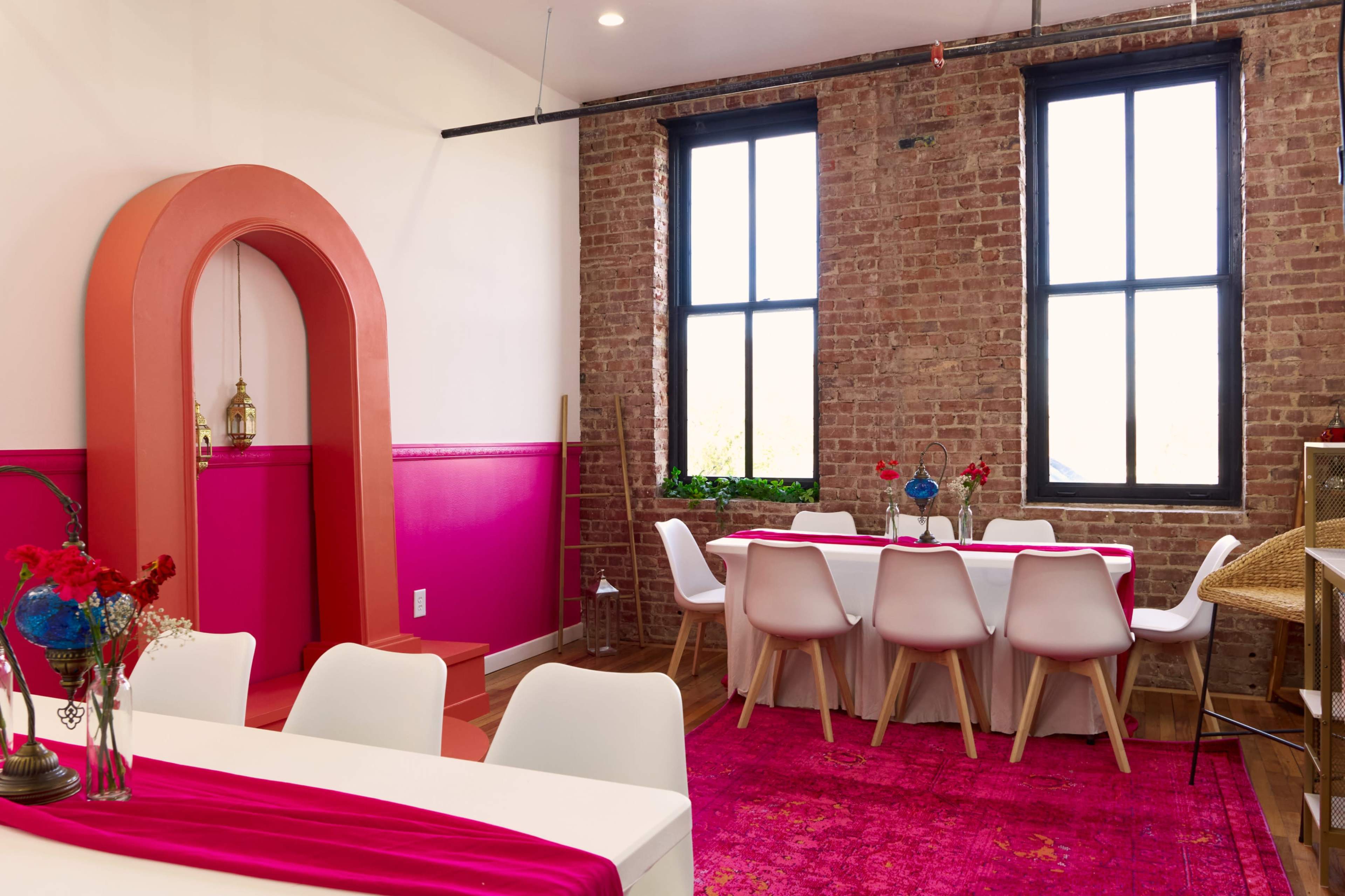 The image shows a bright dining space featuring a white table with chairs, a pink area rug, and exposed brick walls, complemented by large windows.