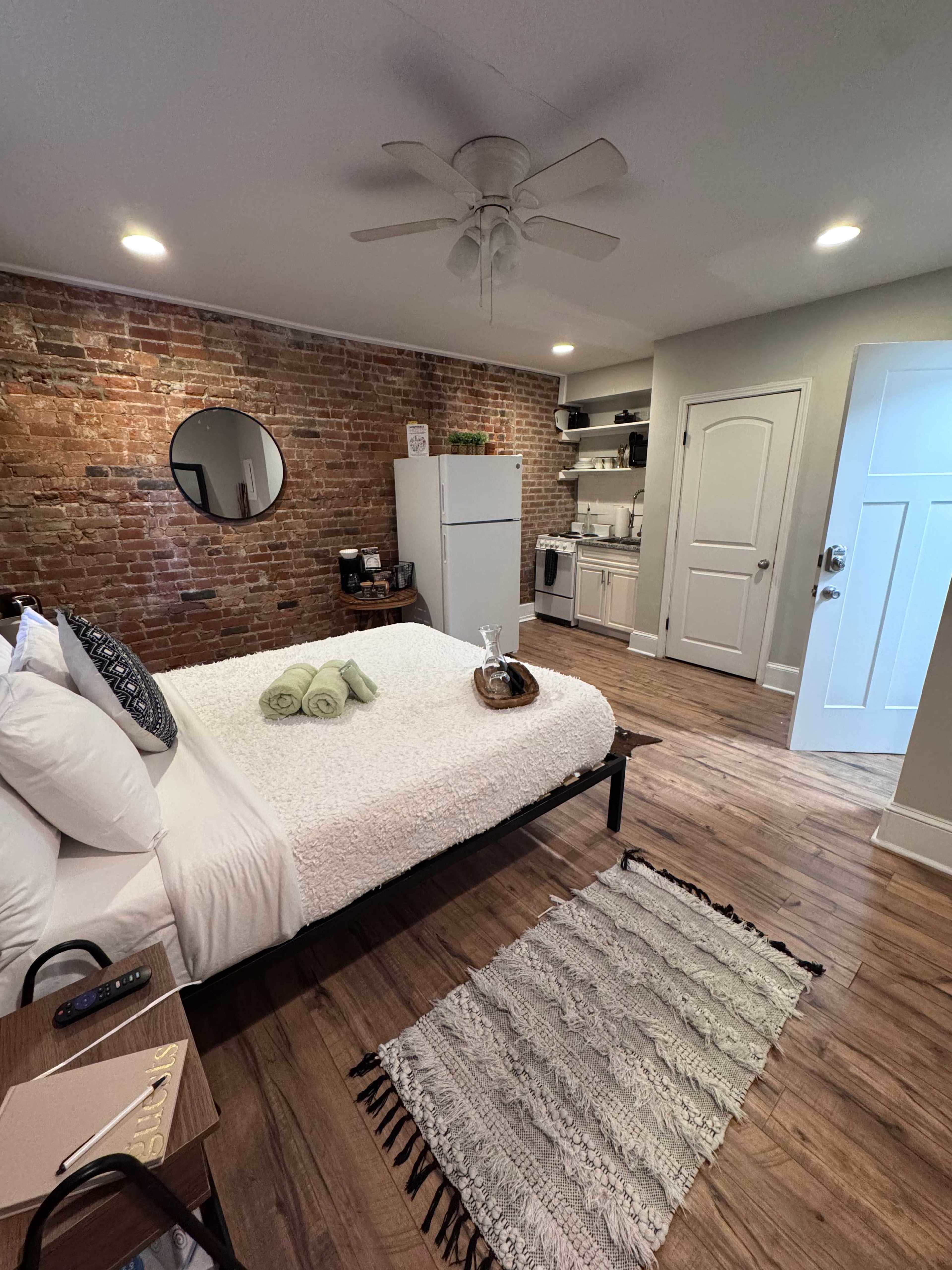 The image shows a compact bedroom with a bed, a small table, a brick accent wall, and a kitchenette area featuring a fridge and shelves.
