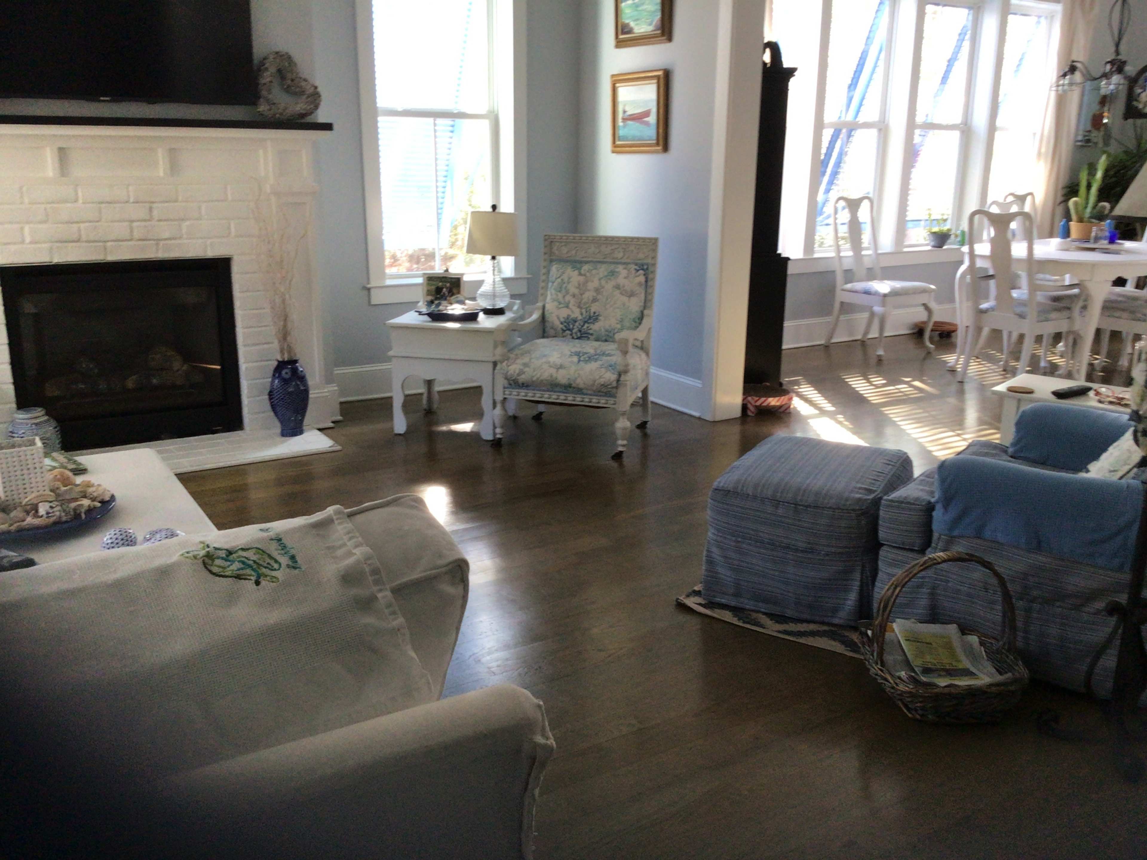 A bright living room with a white brick fireplace, a blue and white patterned armchair, and a dining area visible in the background.