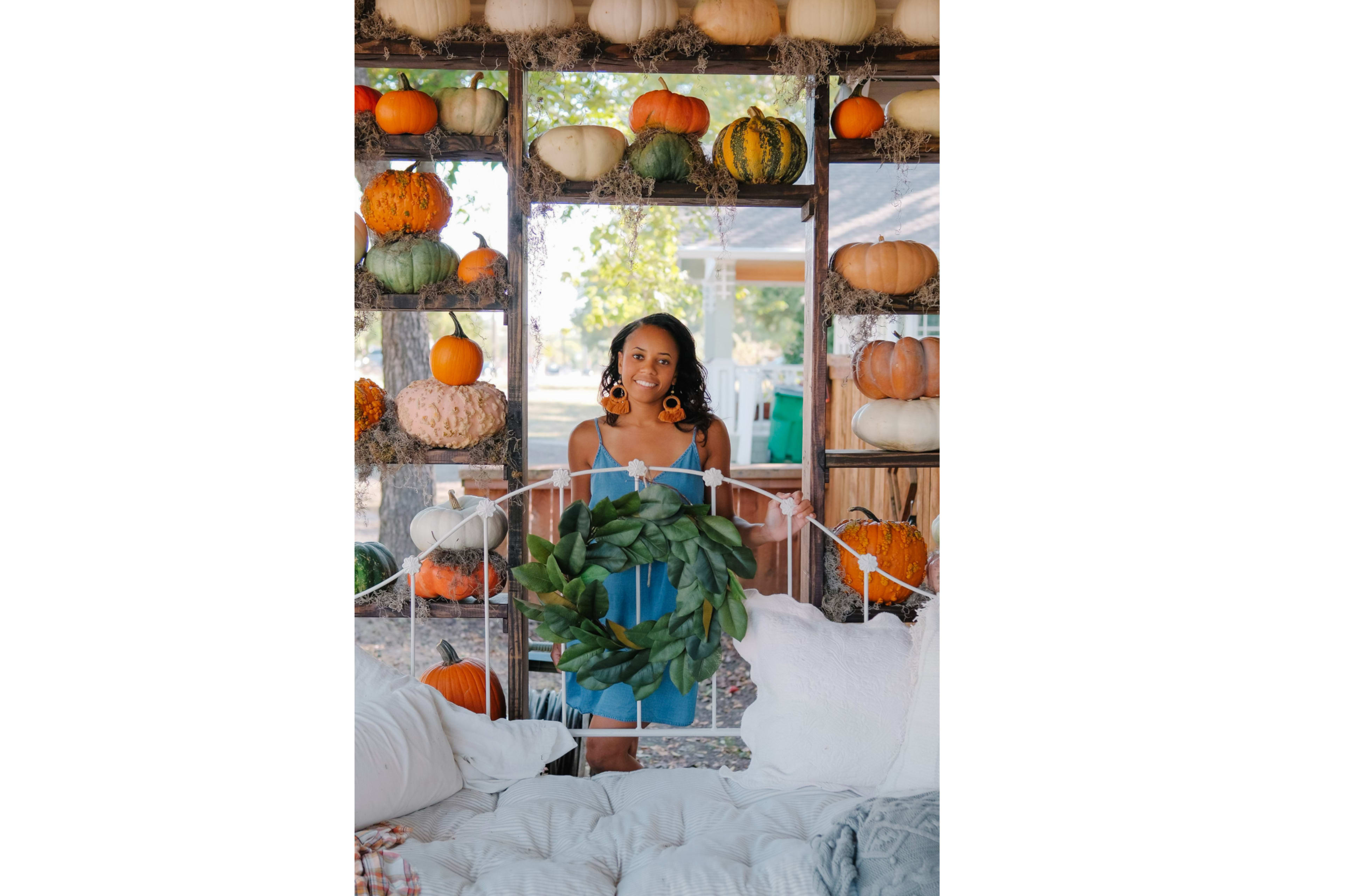 A woman in a blue dress holds a green wreath while standing in front of a rustic display of pumpkins and gourds.
