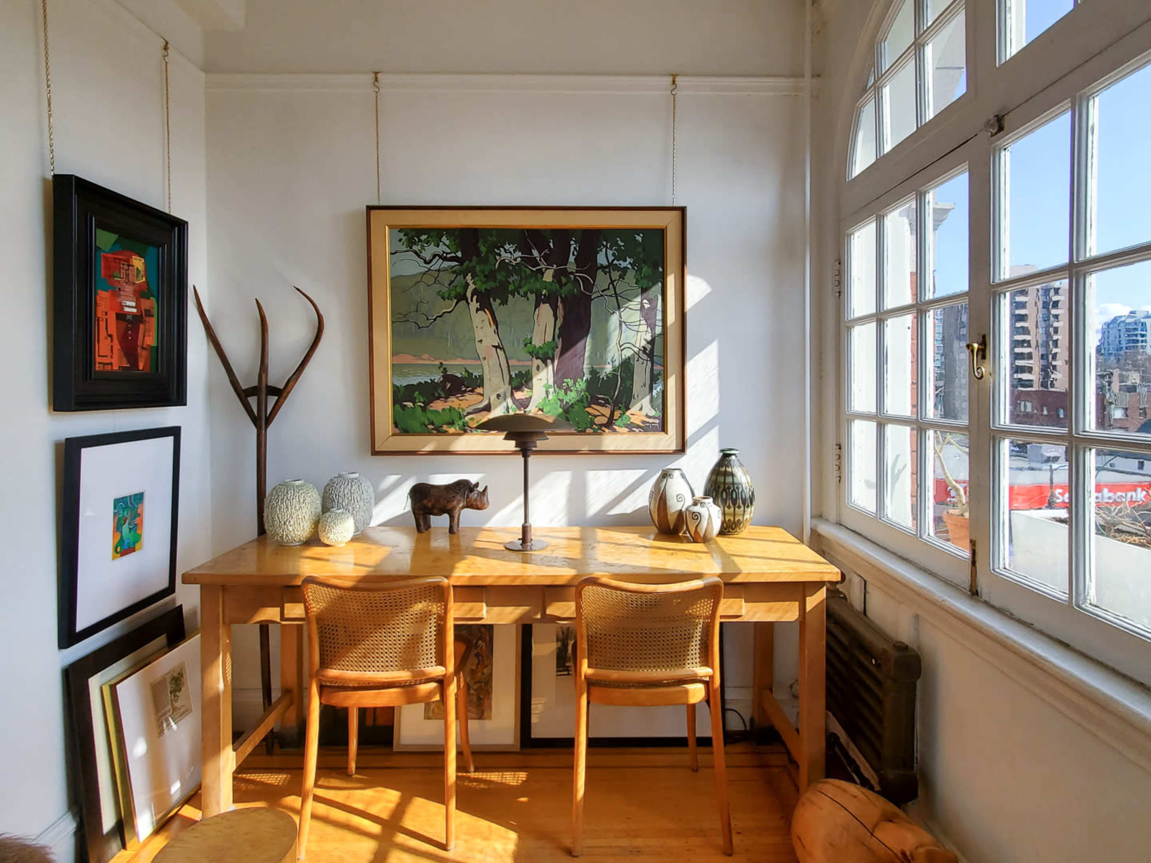 A bright corner of a room features a wooden desk with two chairs, surrounded by artwork and decorative objects, with large windows providing natural light.