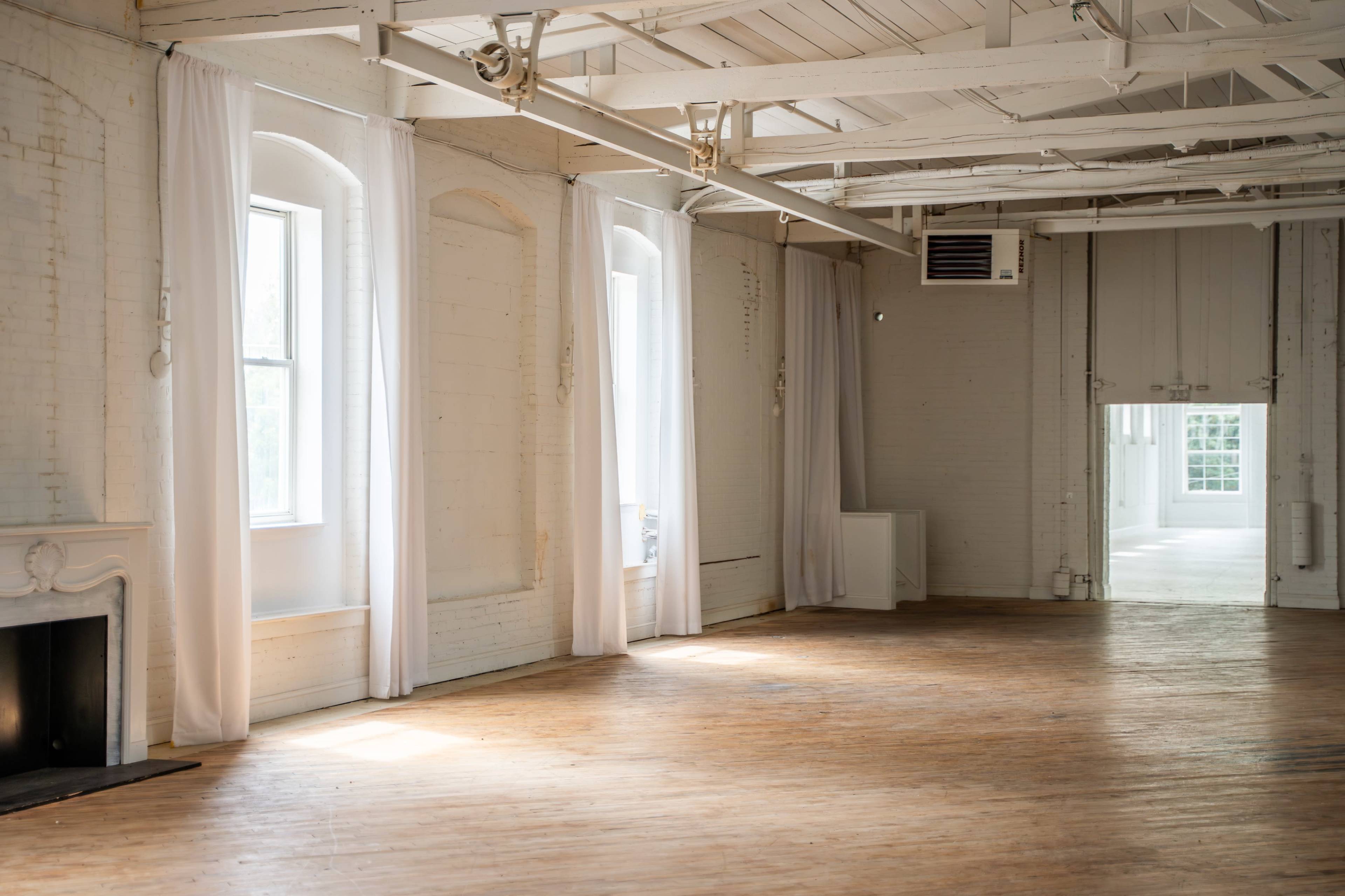 The image shows a spacious, well-lit interior with wooden flooring, large windows draped with white curtains, and a minimalist design.