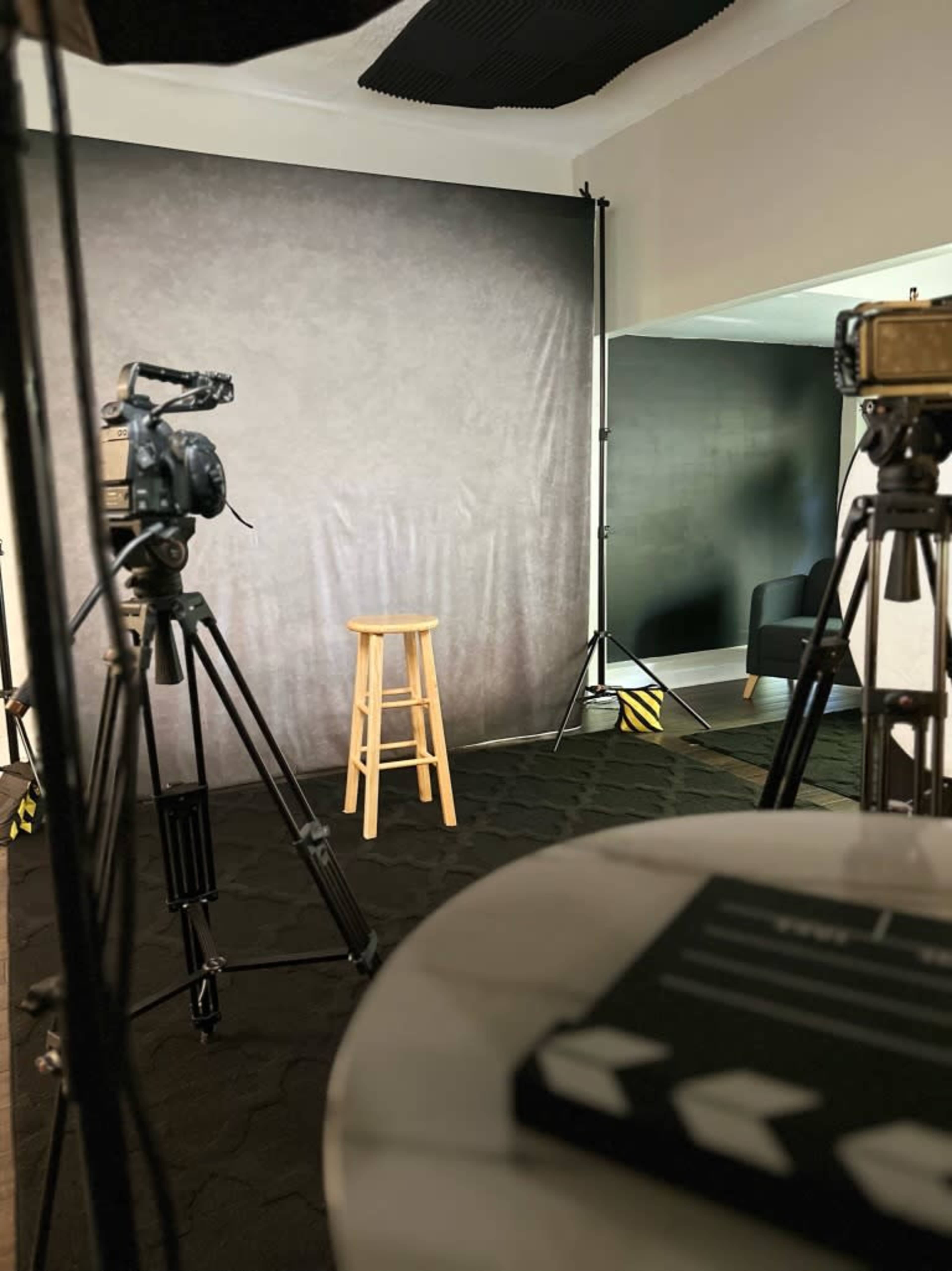 A photography or video recording setup featuring a stool in front of a dark backdrop, with several cameras and tripods positioned around the scene.