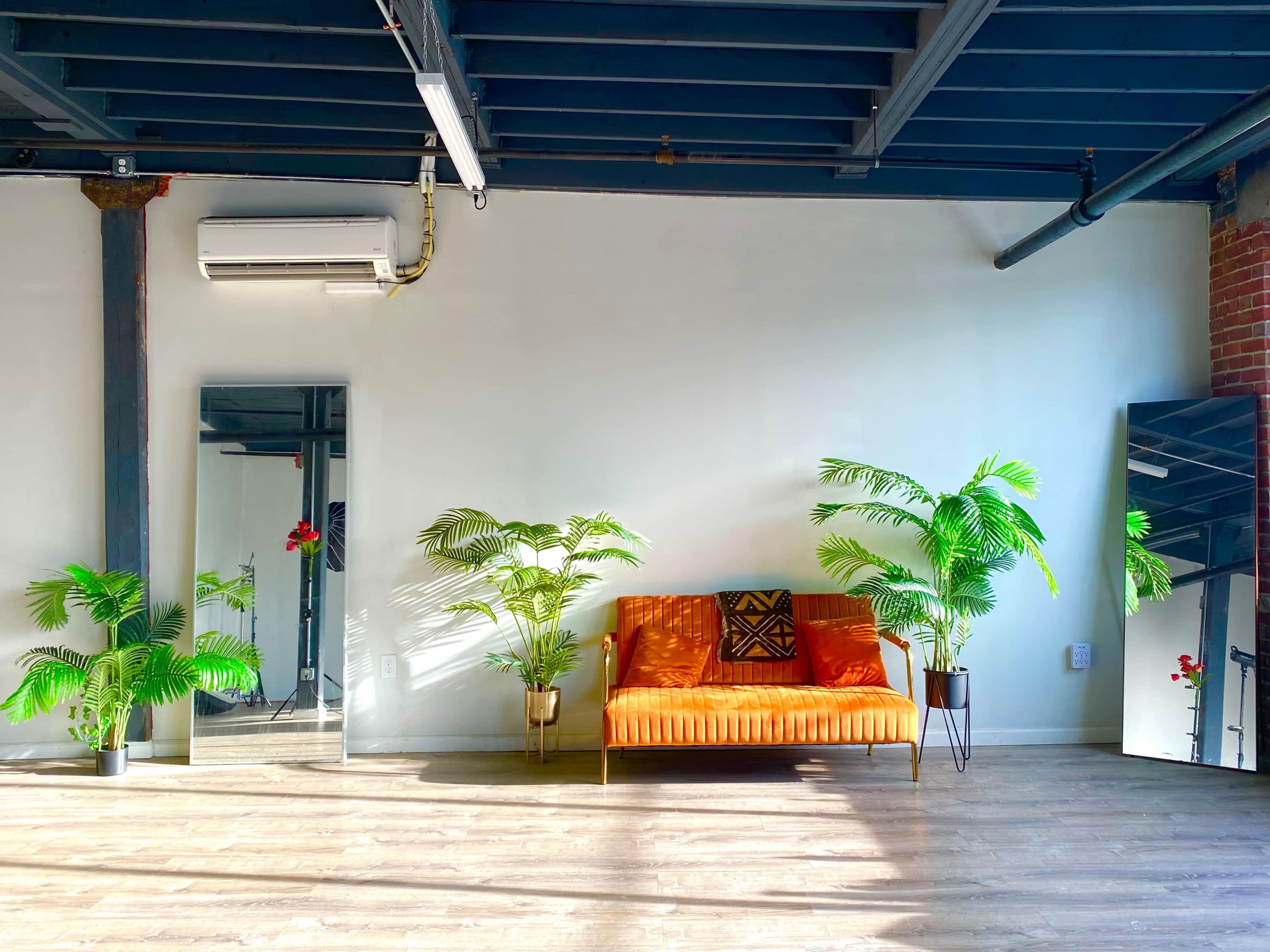 A bright room featuring an orange sofa, a large mirror, and two potted plants on either side.